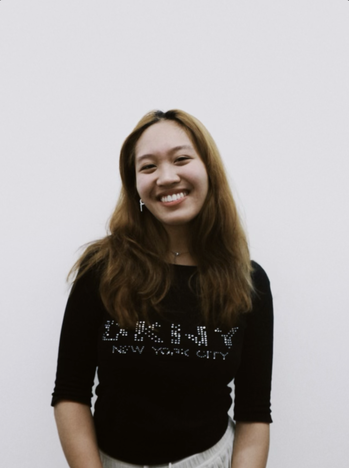 A young woman with light brown hair, smiling, wearing a black shirt with white text and graphics, standing against a plain white background.