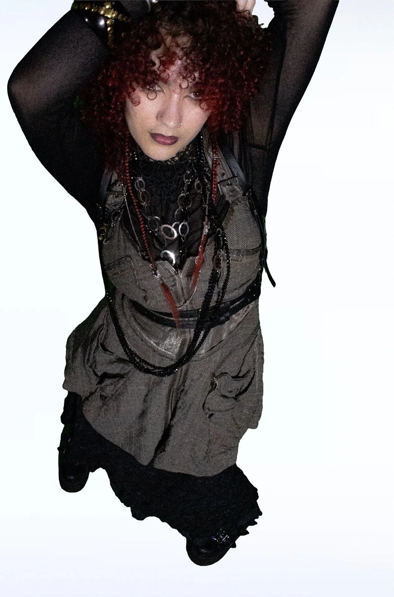 Person with curly red hair posing in black mesh and layered jewelry.
