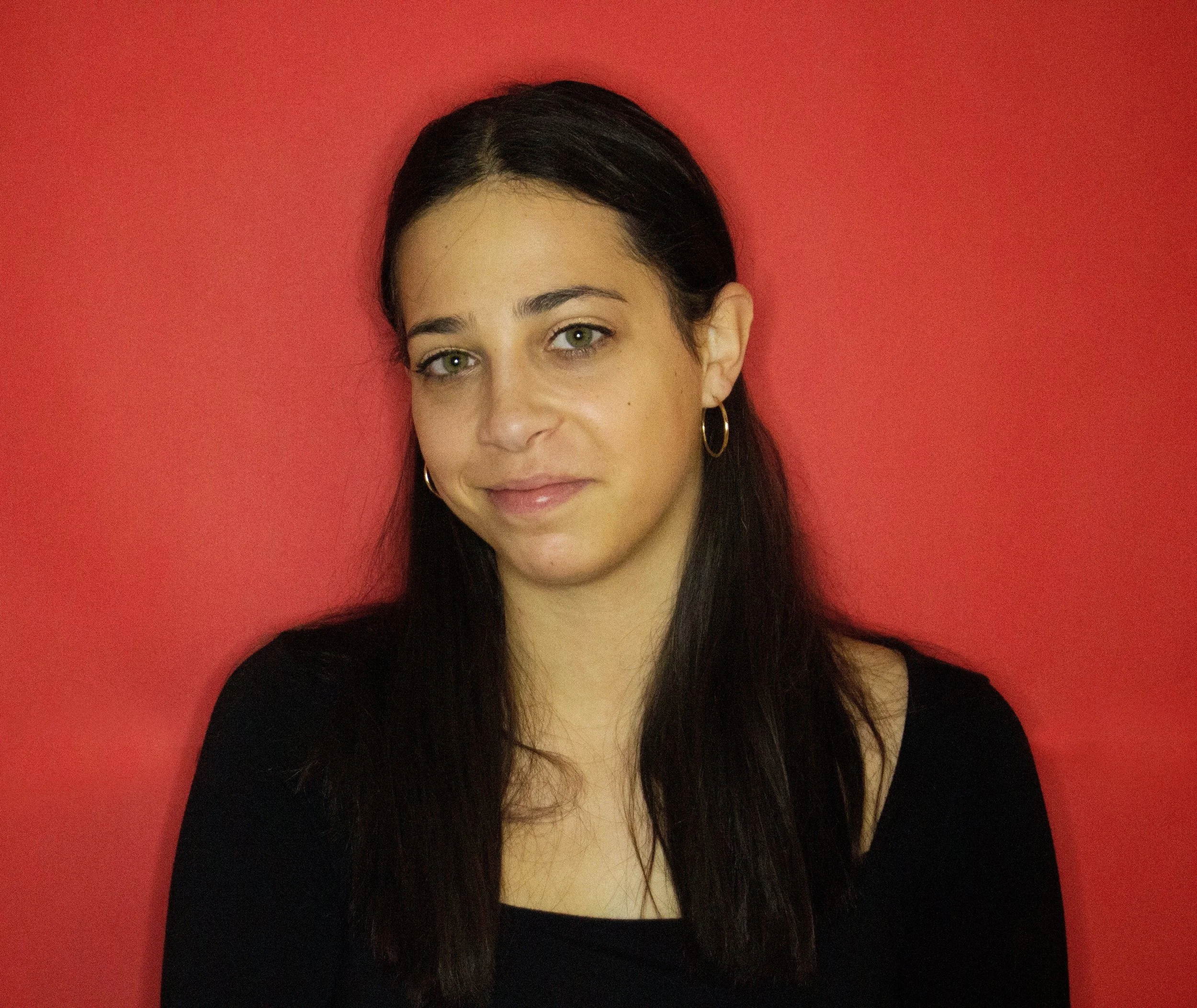 A young woman with dark brown hair and green eyes standing against a red wall, wearing gold hoop earrings and a black top.