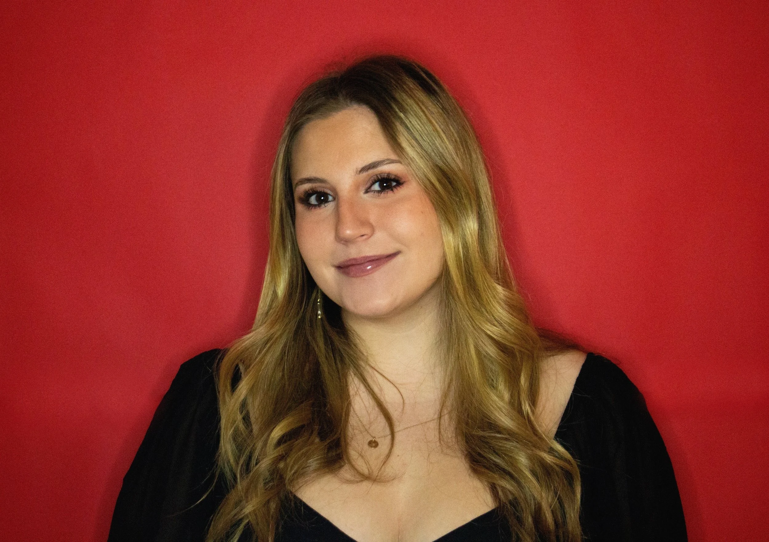 A woman with long, wavy blonde hair, wearing a black top and small hoop earrings, standing against a red background, smiling softly.