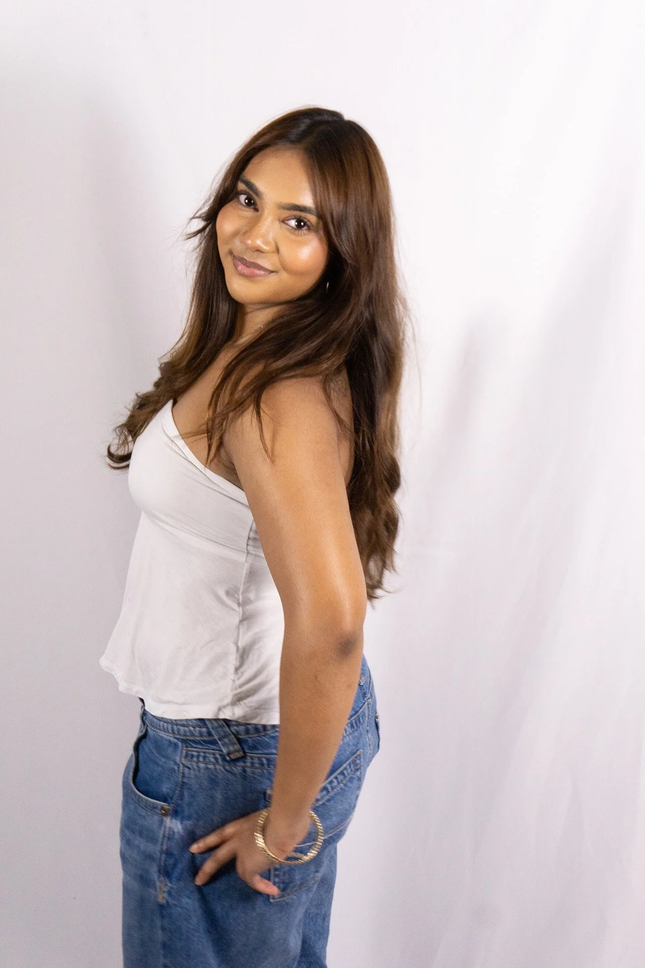 : A South Asian woman stands in a studio against a white backdrop, smiling at the camera while wearing a white top and blue jeans.