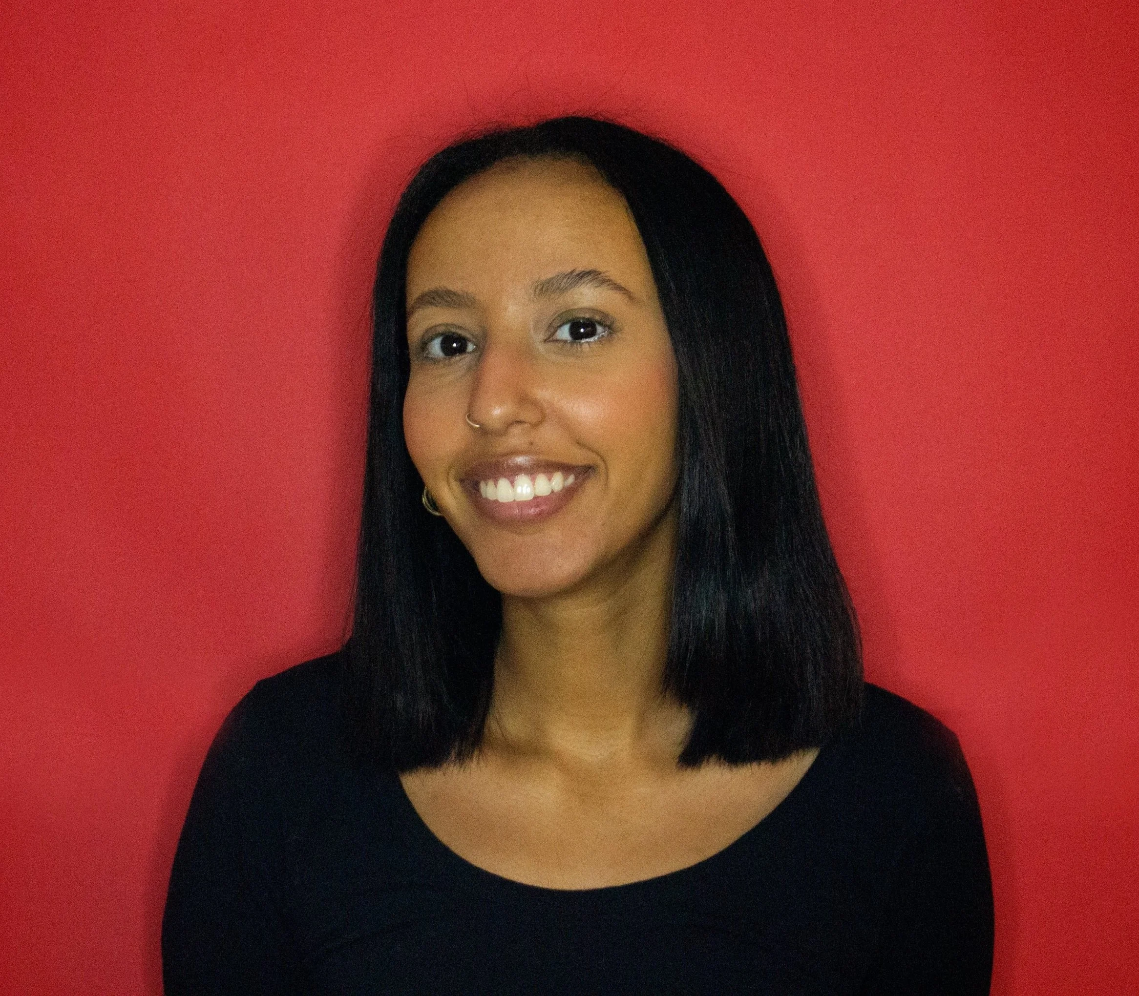 A woman with shoulder-length black hair, a nose ring, and gold hoop earrings, smiling and wearing a black top, standing against a red wall.