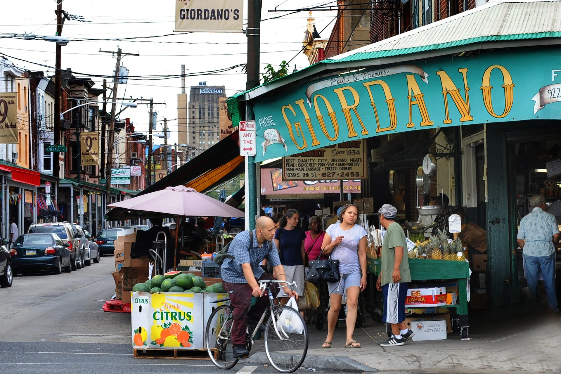 A lively street view of the South 9th Street Italian Market in Philadelphia. The sidewalk is covered by long, green metal awnings that shade a vibrant display of outdoor produce stalls.
