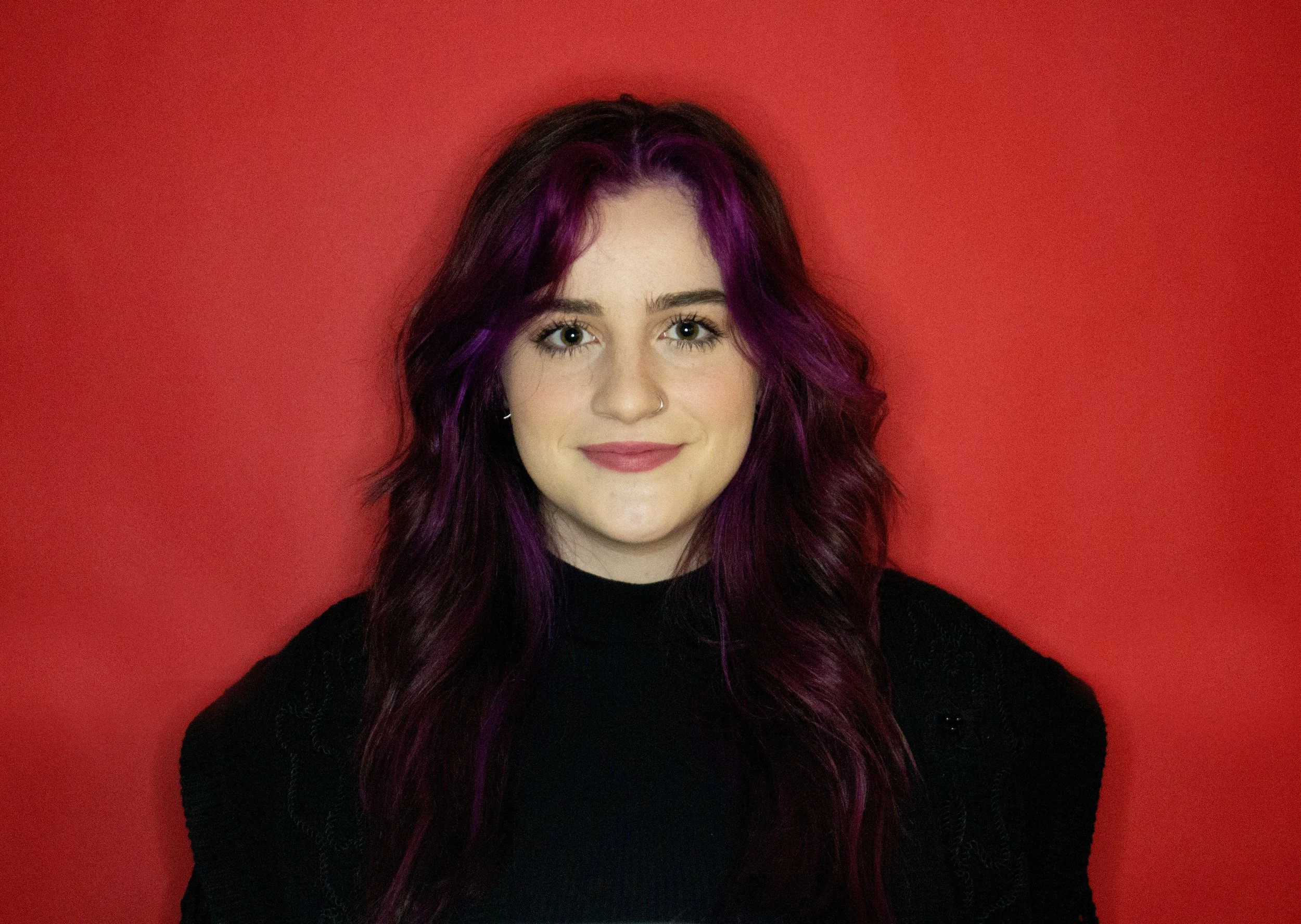 A young woman with long, wavy purple hair, wearing a black top, smiling, standing against a red background.