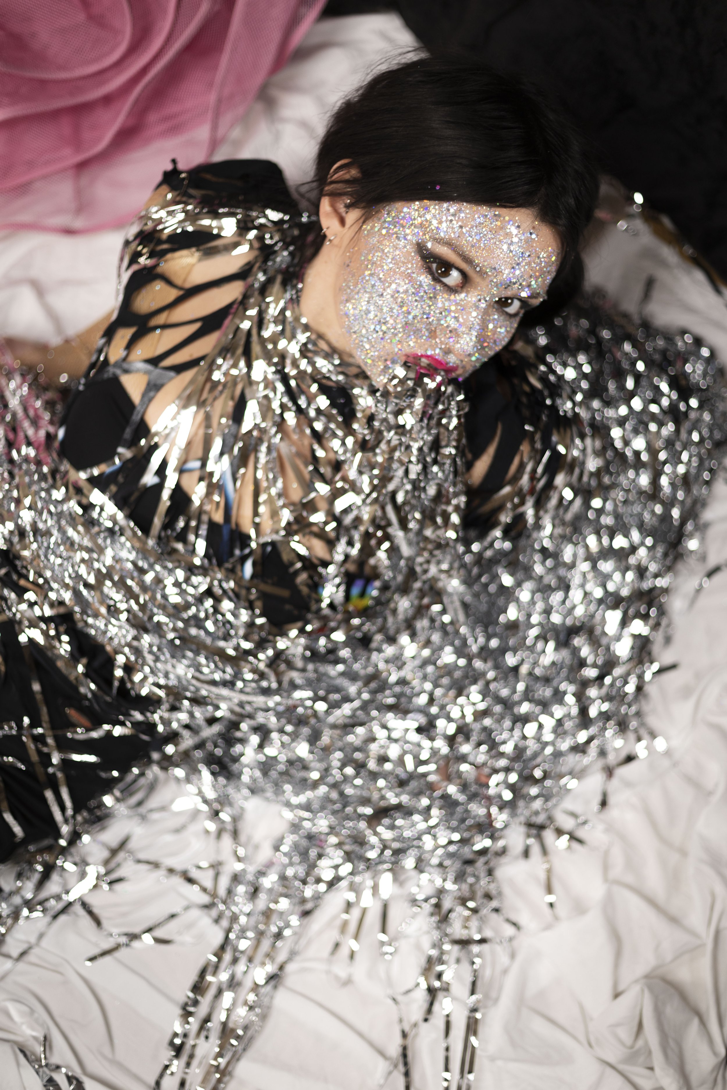 Model with glitter-covered face, wrapped in silver tinsel and wearing a black cutout outfit, looking directly at the camera.