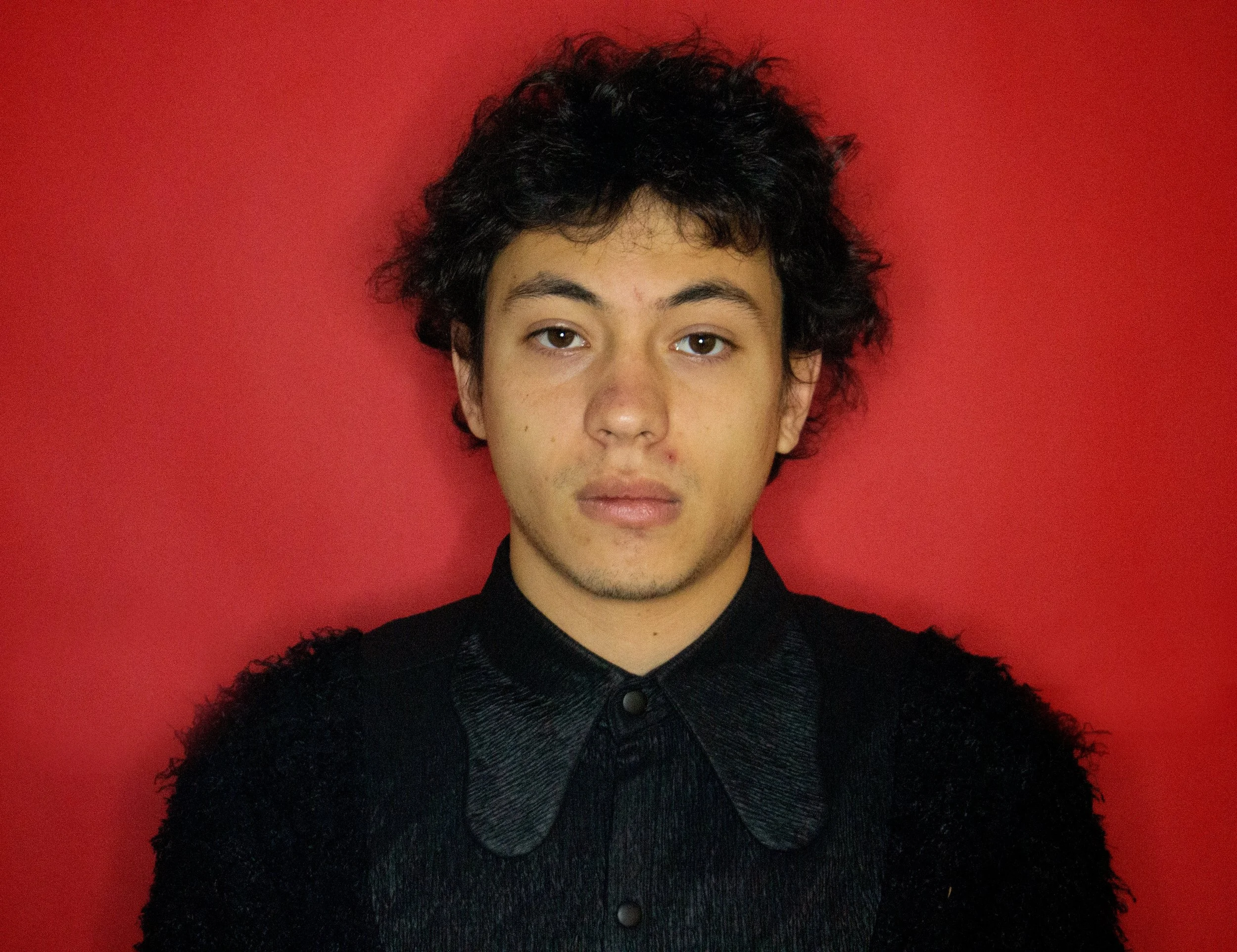 Young man with curly black hair wearing a black shirt in front of a red background.