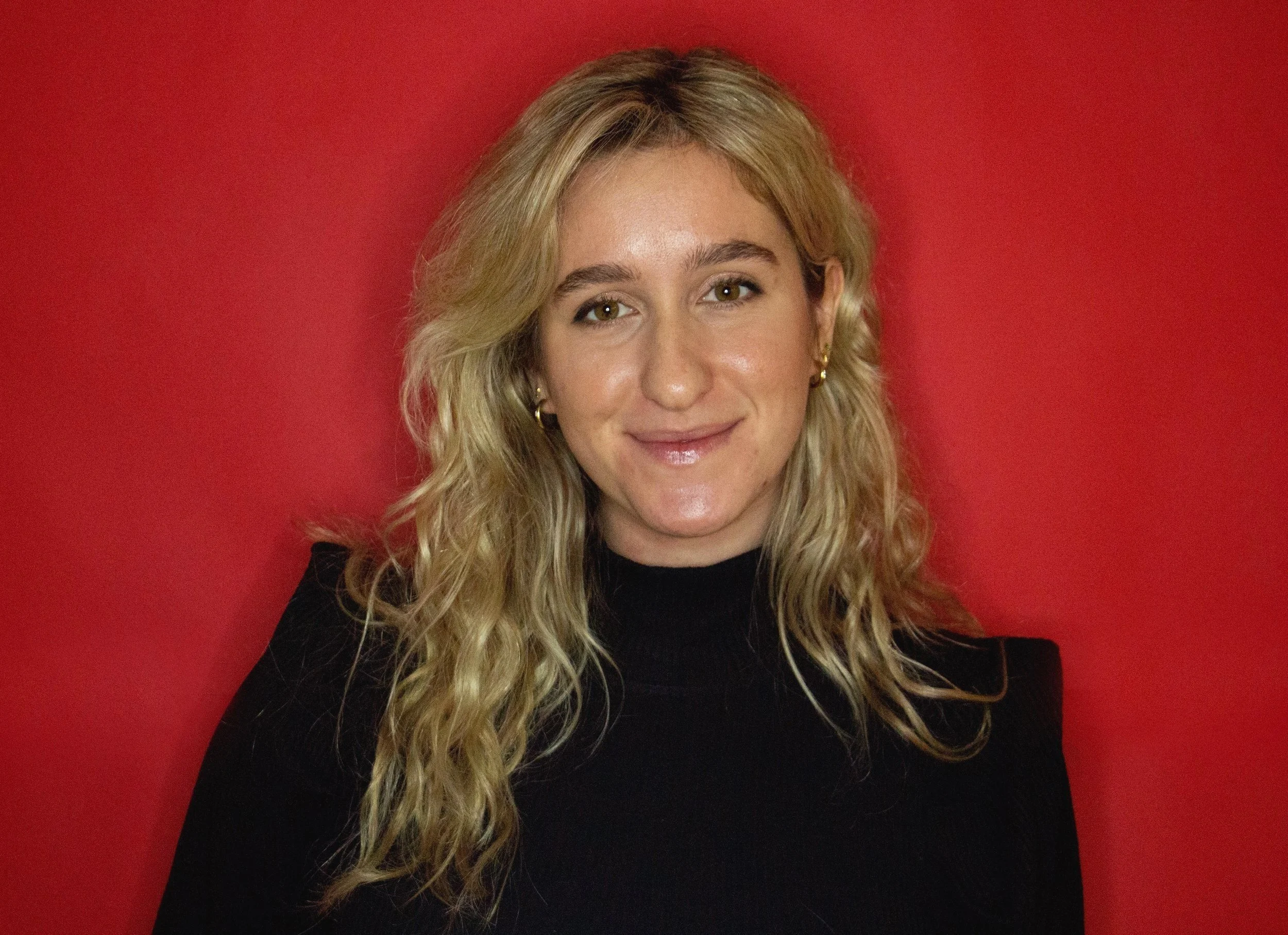 A woman with blonde curly hair, wearing a black top and small hoop earrings, smiling against a red background.