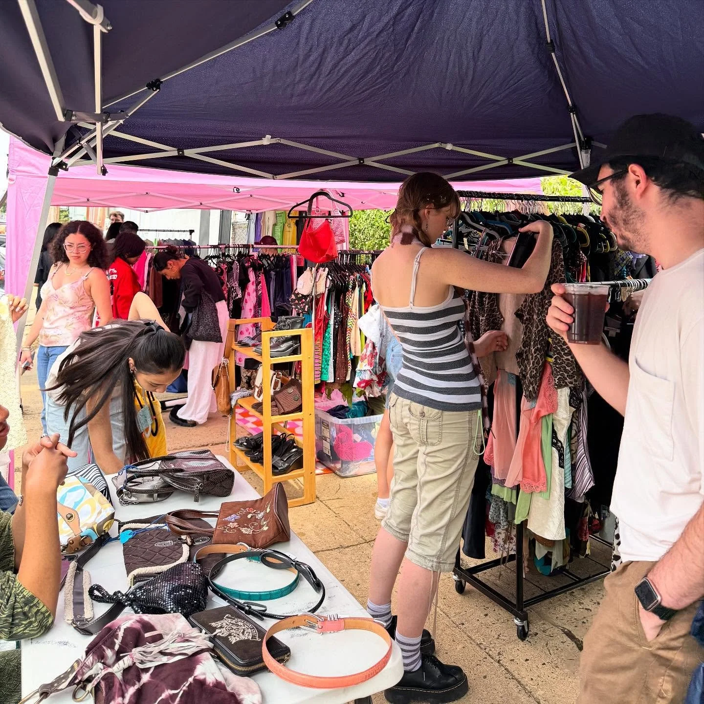 A bustling outdoor flea market scene on a sunny day. White pop-up tents line a paved area, filled with a curated selection of vintage furniture, handmade crafts, and antique home decor.