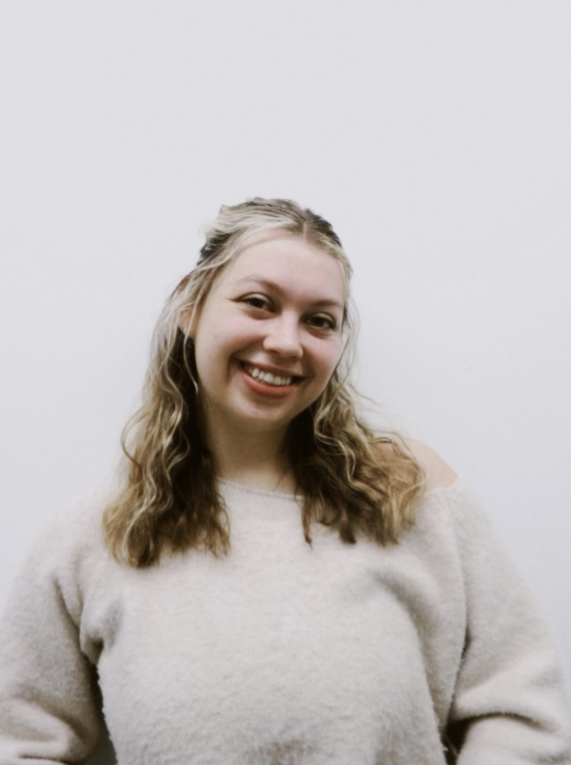 A smiling woman with wavy blonde hair wearing a beige sweater standing against a plain white wall.