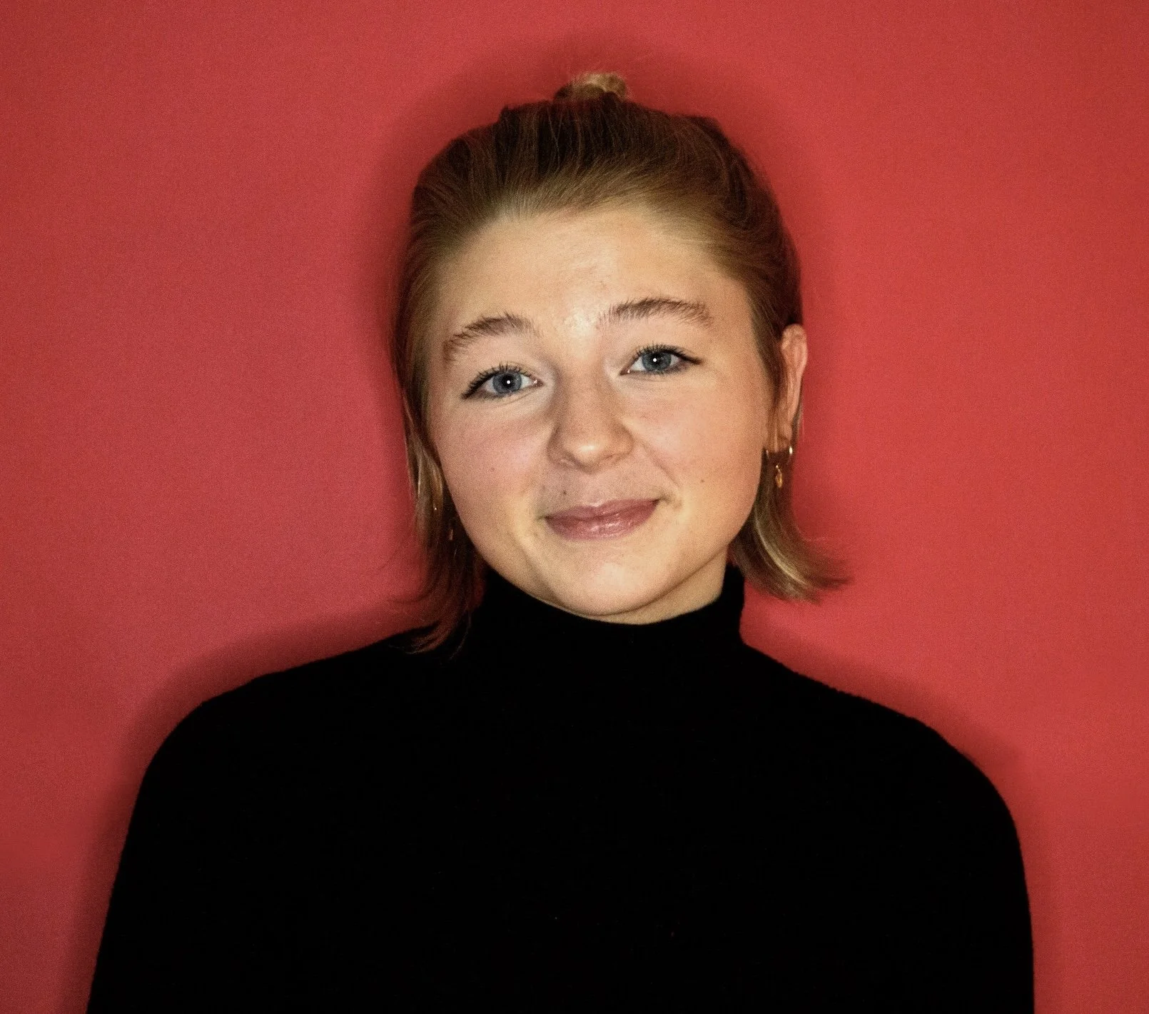 A young woman with short blonde hair, blue eyes, wearing a black turtleneck sweater, smiling, standing against a red background.