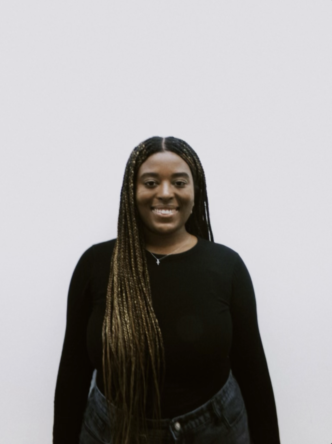 A young woman with long braided hair, wearing a black long-sleeve shirt and dark jeans, smiling in front of a plain white wall.