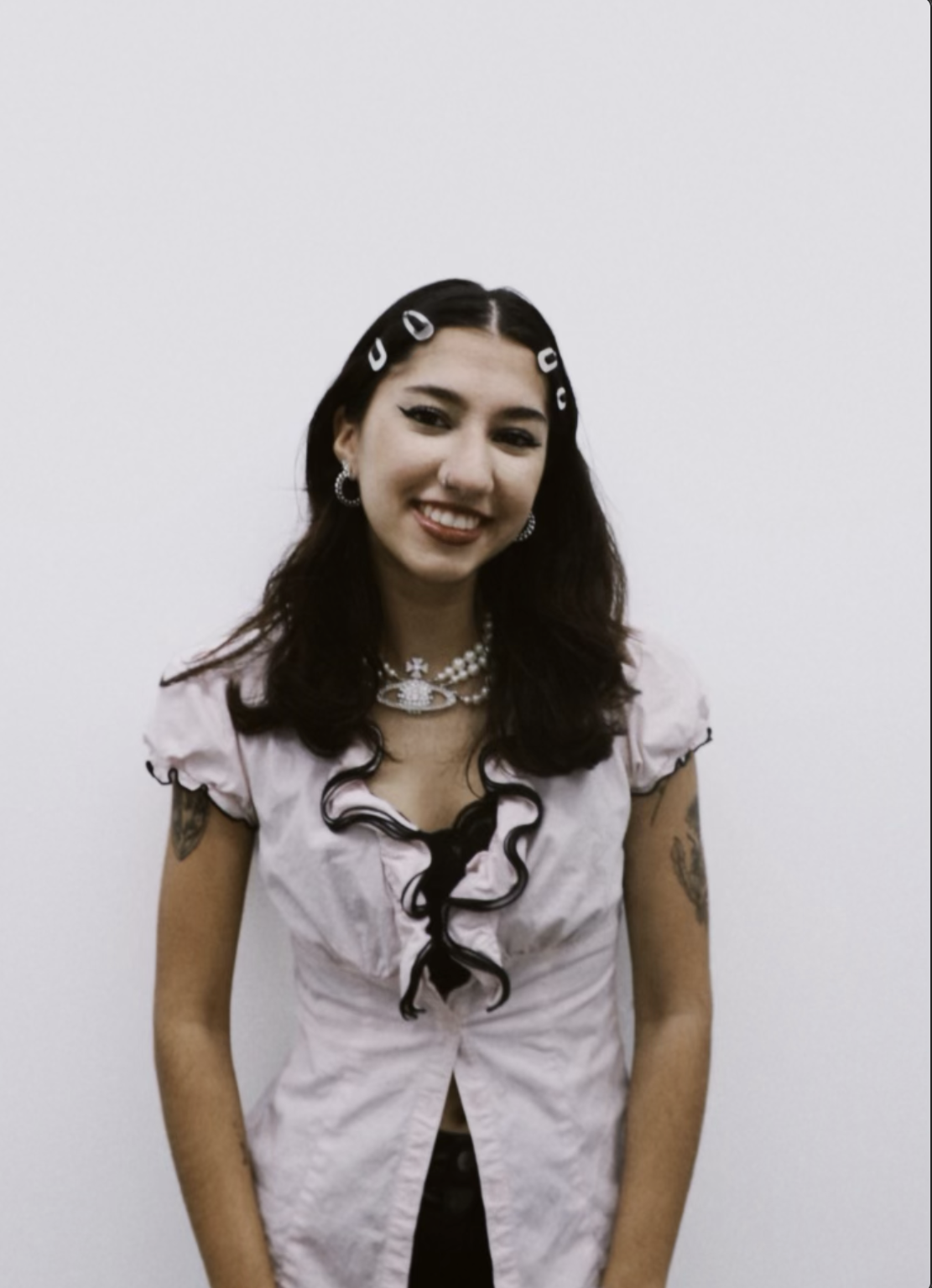 Young woman with dark hair, wearing pink top with black curls, earrings, necklaces, smiling, standing against a plain white wall.