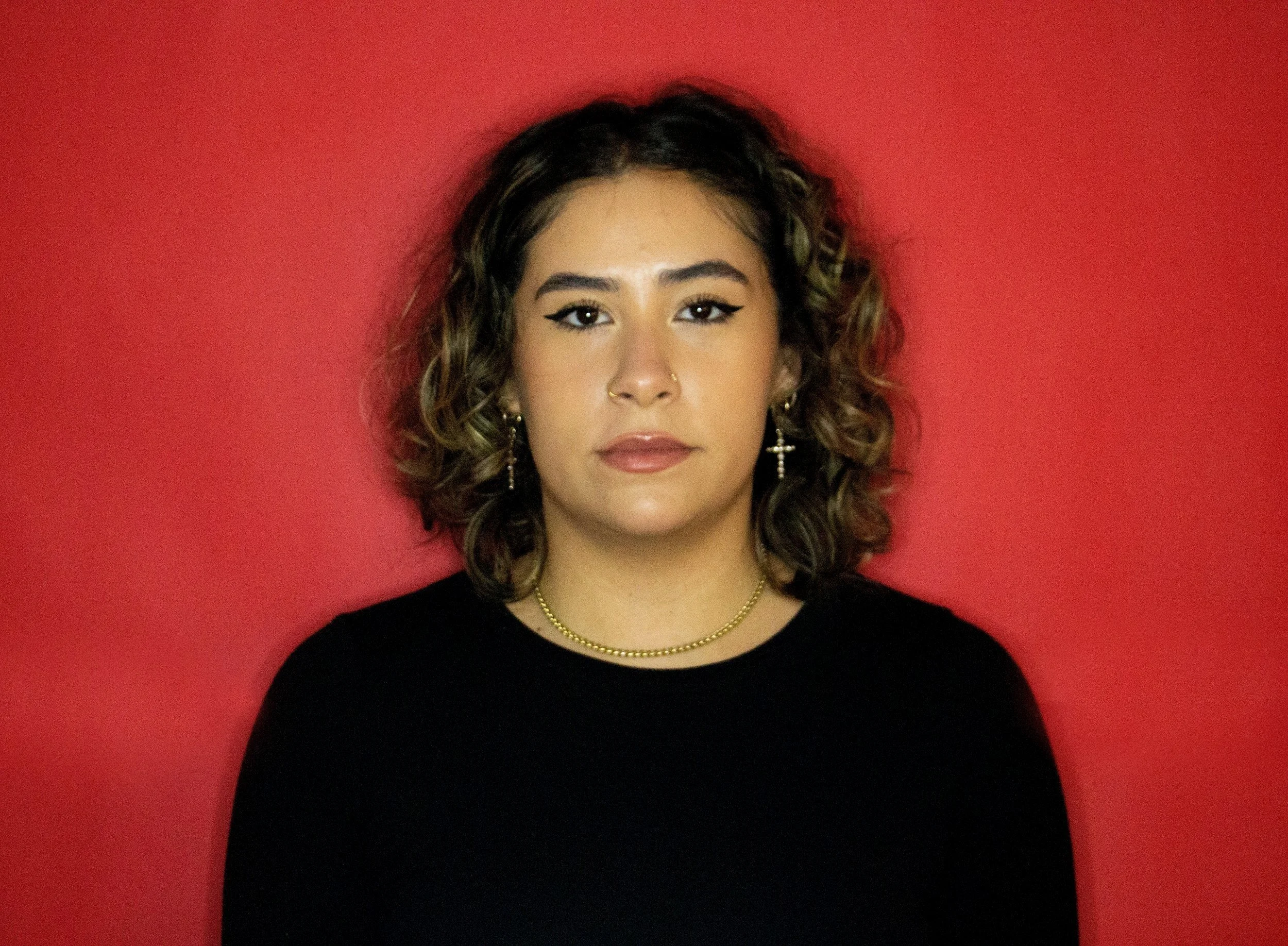 A woman with medium-length curly hair, wearing a black top, gold earrings, and a gold necklace, posed against a solid red background.