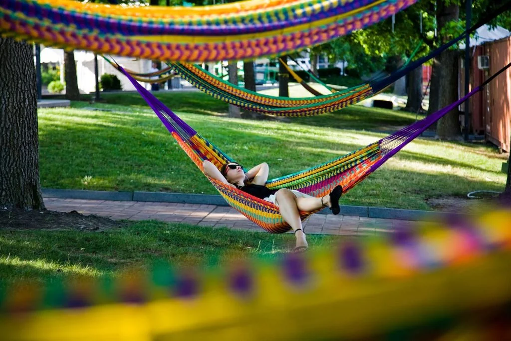 A festive nighttime scene at an urban waterfront park. Dozens of colorful hammocks in shades of pink, blue, and yellow are strung between mature trees.