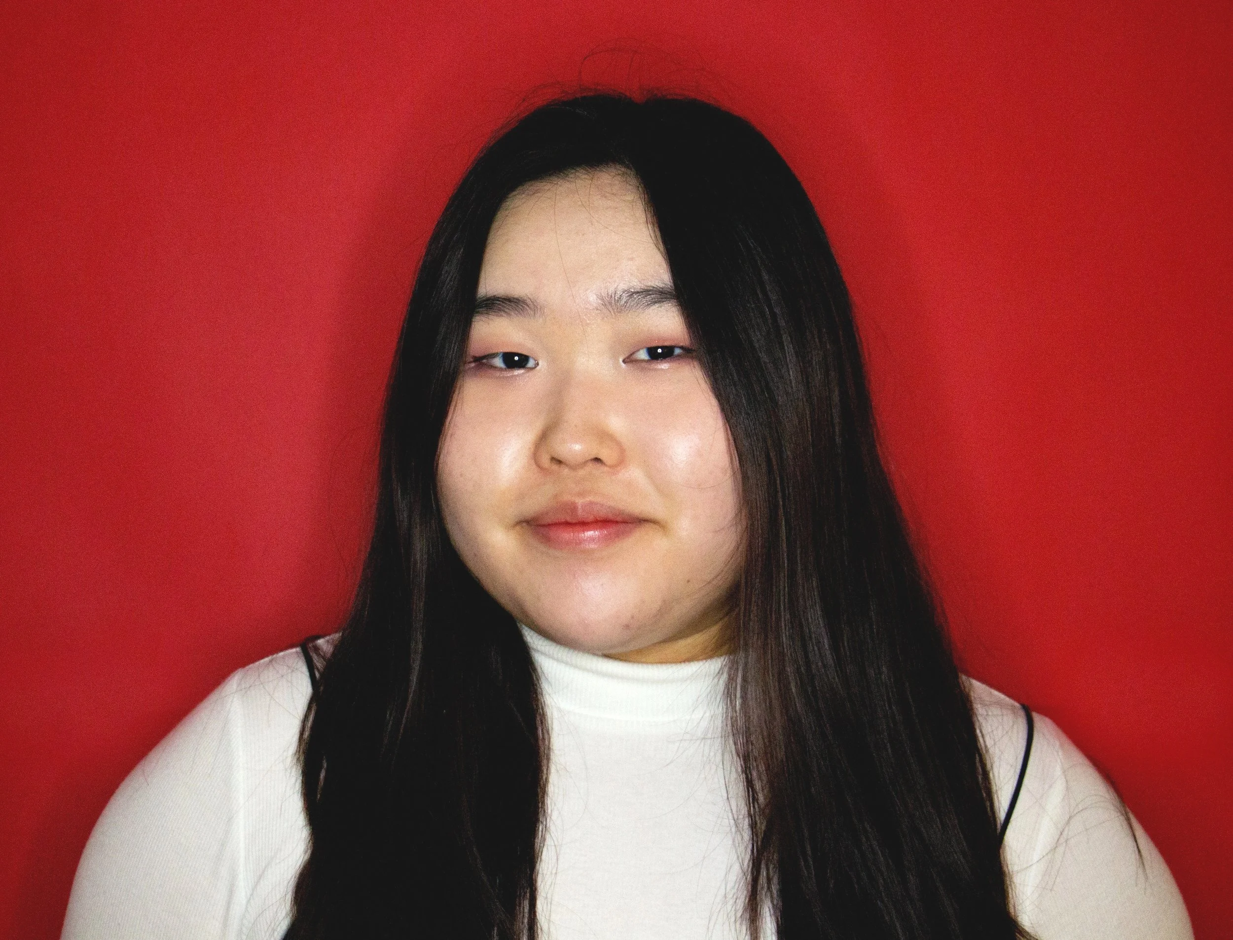 A young woman with long black hair wearing a white turtleneck top posing against a red background.