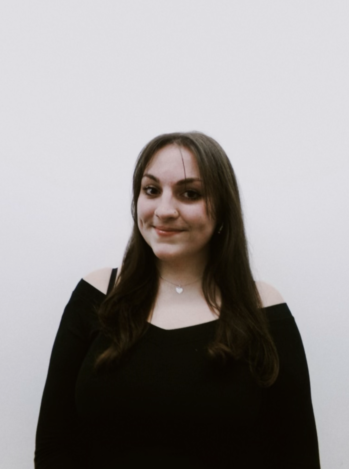 A young woman with long brown hair smiling at the camera, wearing a black cold shoulder top and a silver heart necklace was a white background.