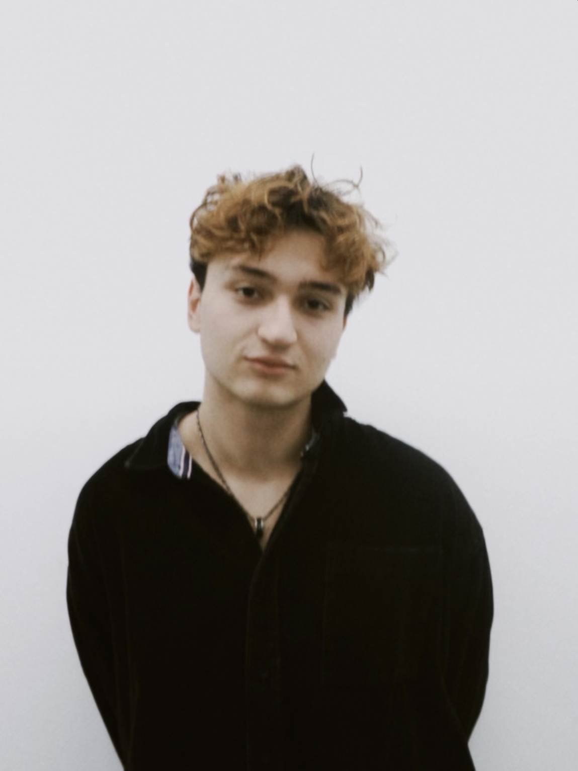 A young man with curly, reddish hair wearing a black jacket and a necklace stands against a plain white background.
