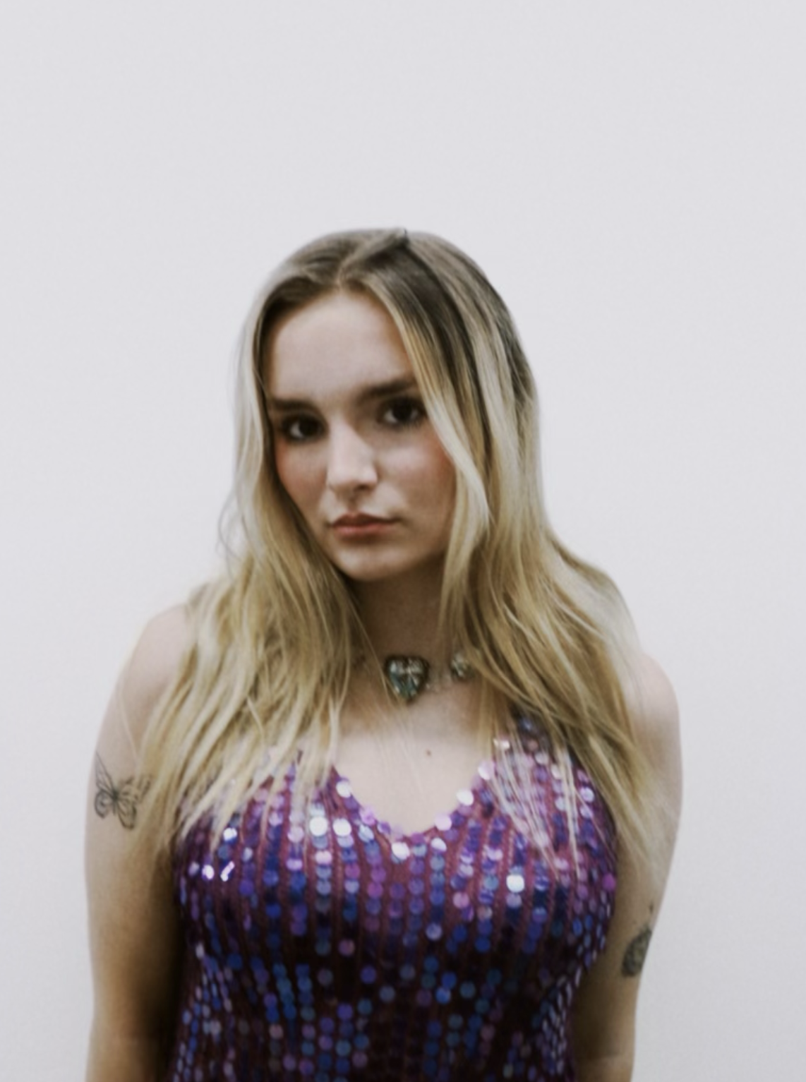 Young woman with long blonde hair wearing a purple sequined top and a necklace, standing against a plain white background.