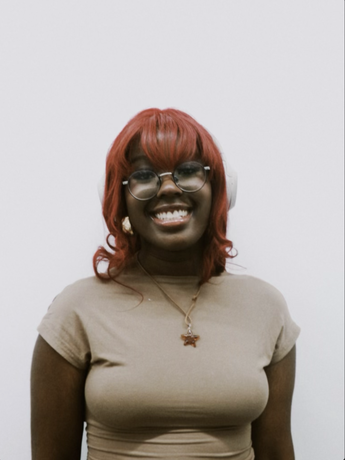 A woman with red hair, glasses, and headphones, smiling at the camera, wearing a beige t-shirt and a necklace with a pendant.