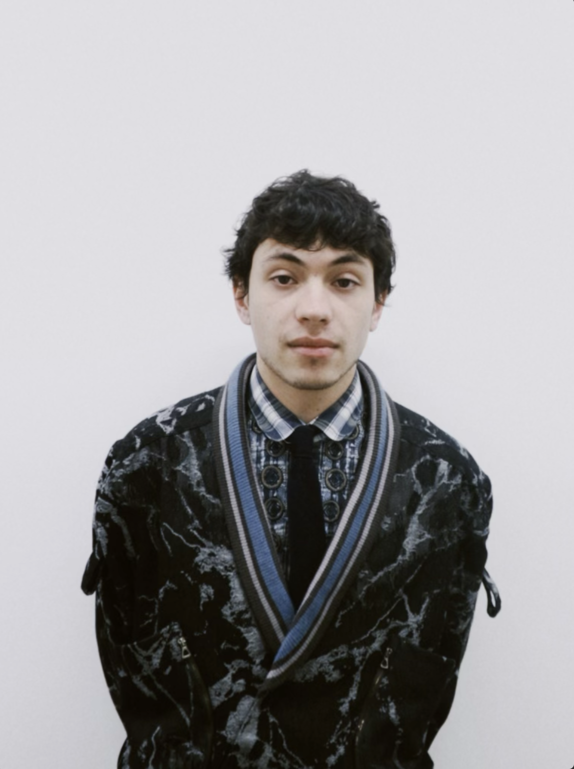 Young man with dark wavy hair wearing a patterned black and white jacket, plaid shirt, and tie, standing against a plain white background.