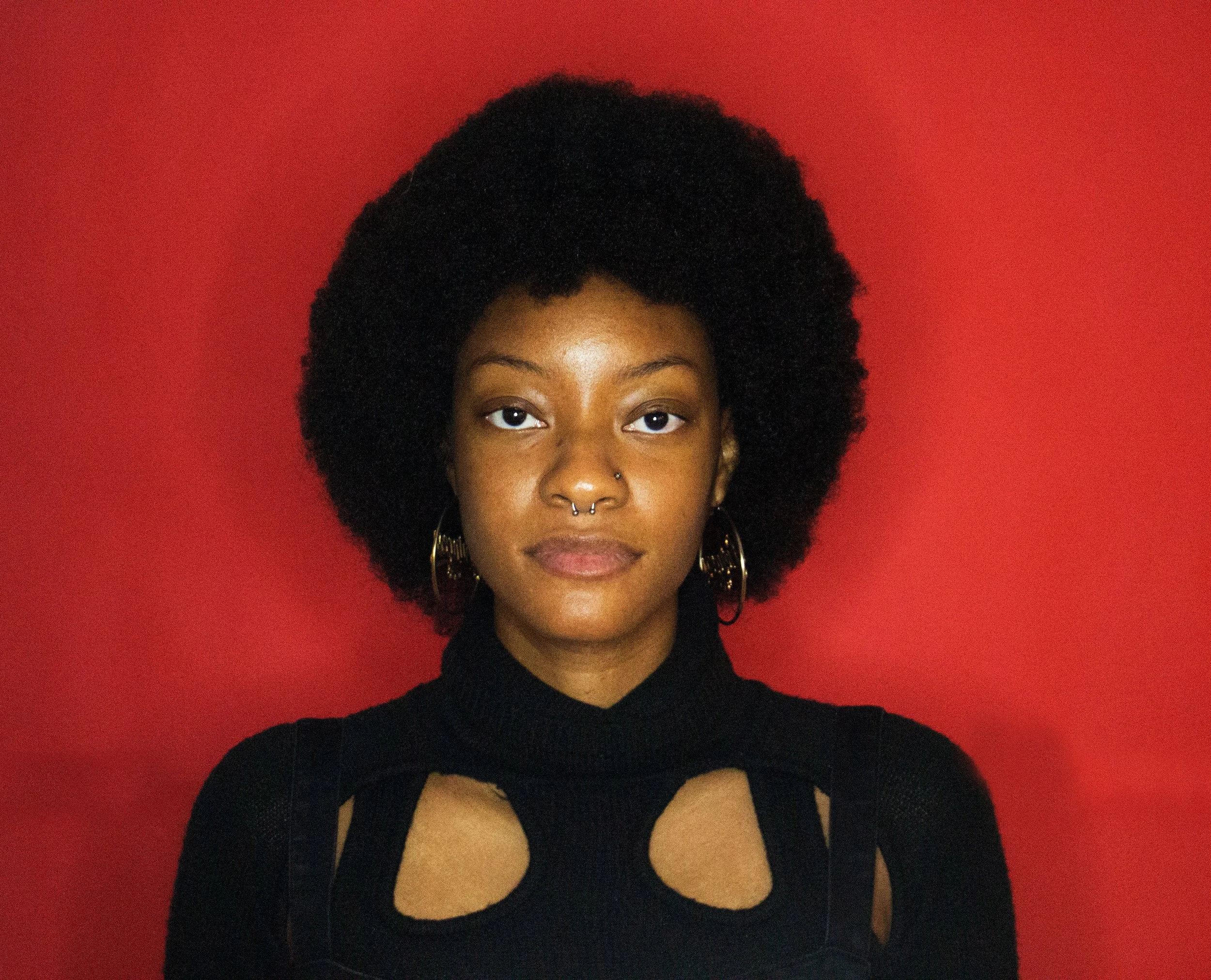 A young African American woman with a natural afro hairstyle, wearing a black top with cut-out details on the shoulders, gold hoop earrings, and has a septum piercing, standing against a red background.