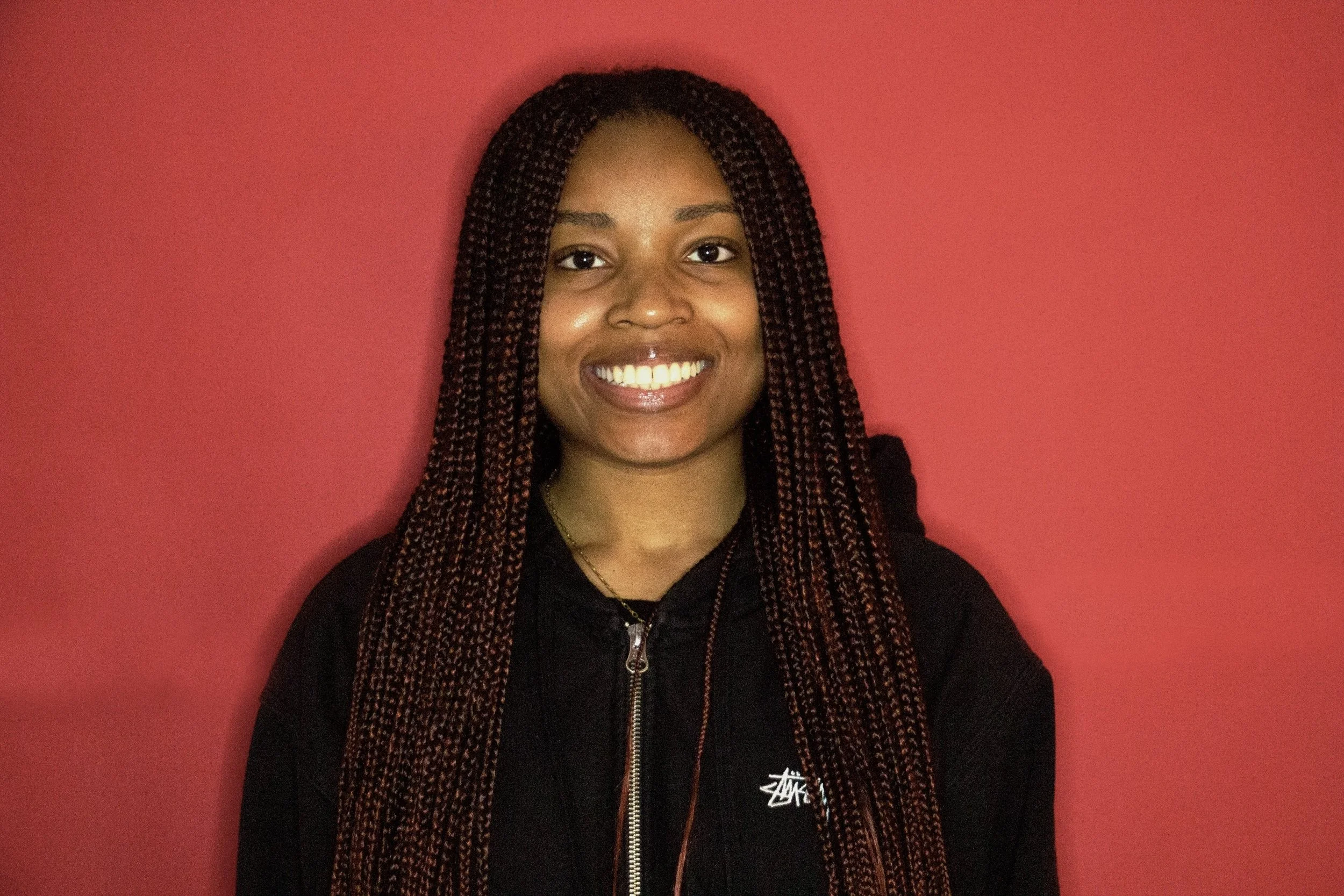 A young woman with long braids smiling in front of a pink wall.