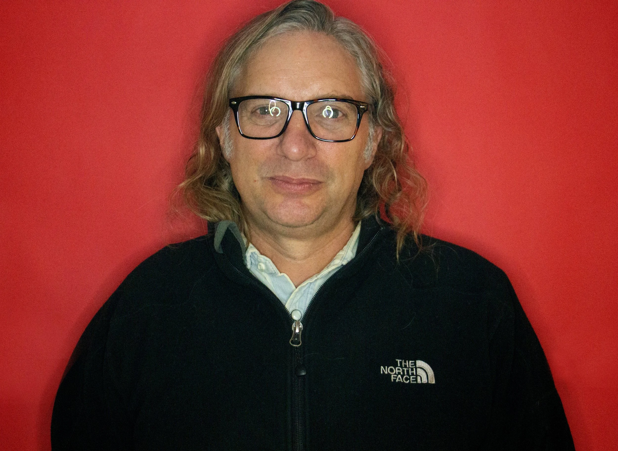A man with shoulder-length blonde hair, glasses, and a light beard, wearing a black North Face jacket and a white shirt, standing against a red background.