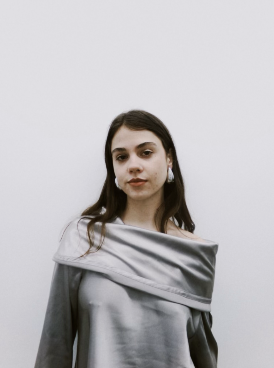 Young woman with long dark hair wearing silver satin off-shoulder dress and earrings, posing against a plain white wall.