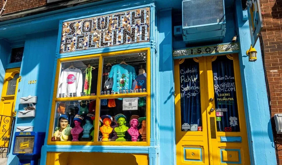 The quirky and colorful storefront of South Fellini on East Passyunk Avenue. The window display is packed with hyper-local Philadelphia "inside joke" merchandise