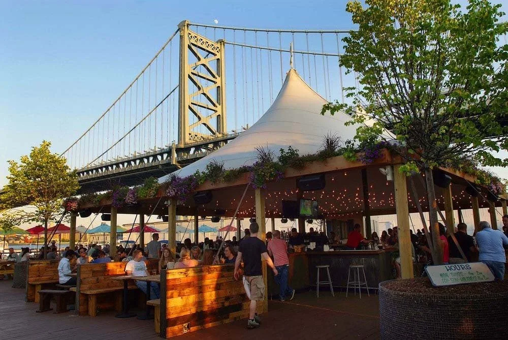 A wide-angle view of an expansive outdoor waterfront beer garden located on the Delaware River. Rows of wooden picnic tables and yellow chairs are arranged under strings of Edison lights and large wooden umbrellas.
