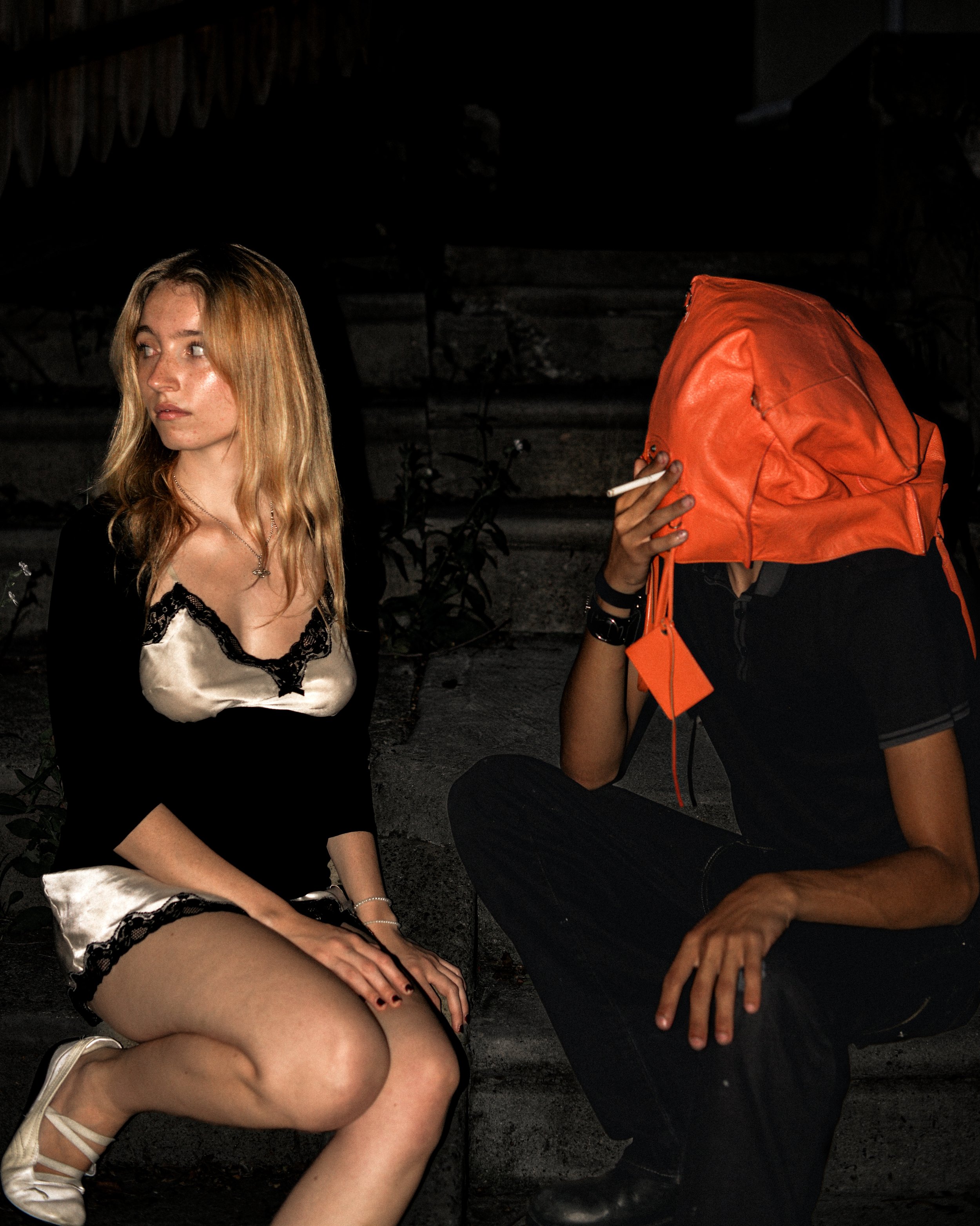 A woman sits beside a figure wearing an orange Balenciaga bag over his head on outdoor steps at night.