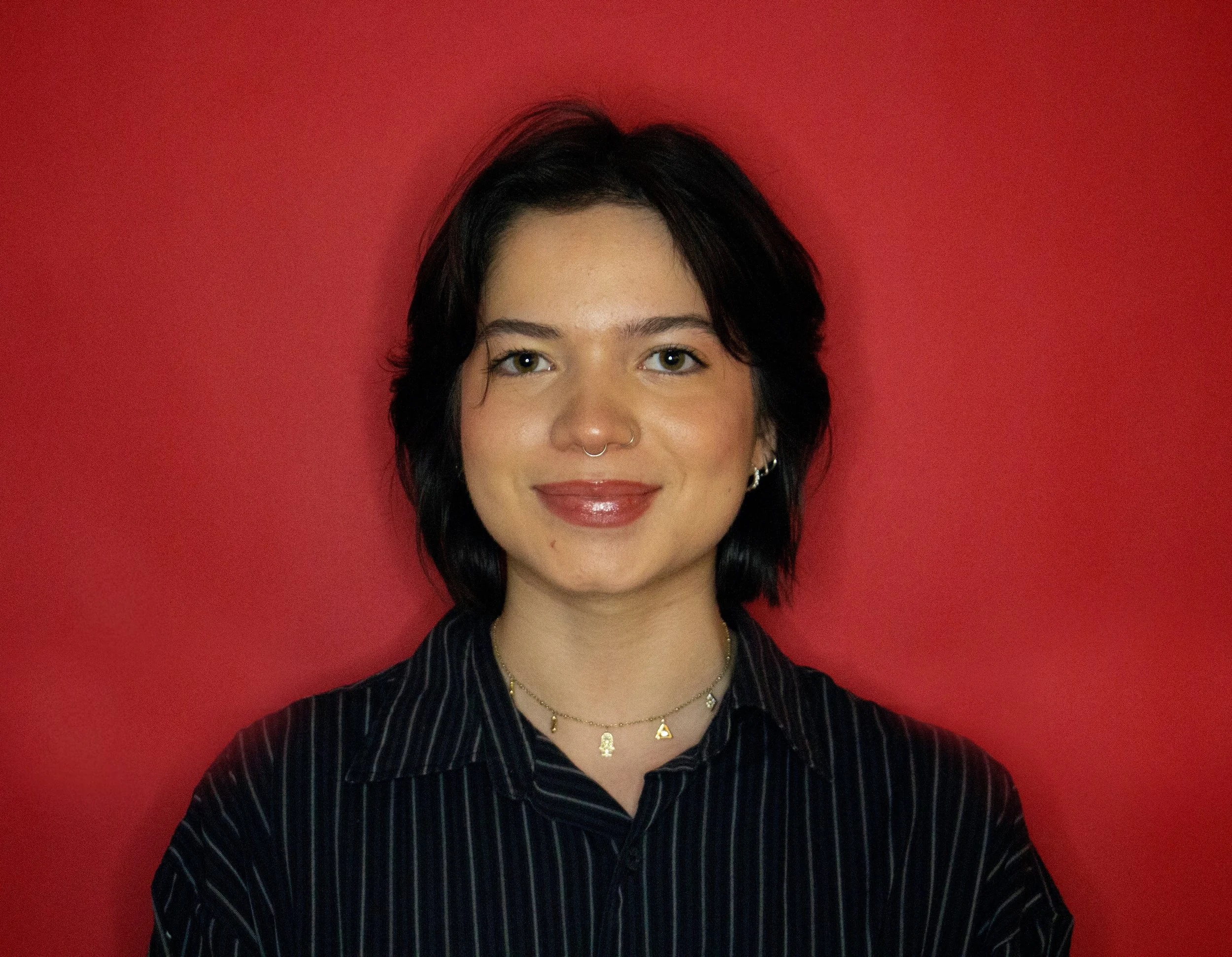 A young woman with short black hair, wearing a black striped shirt, smiling against a red background.