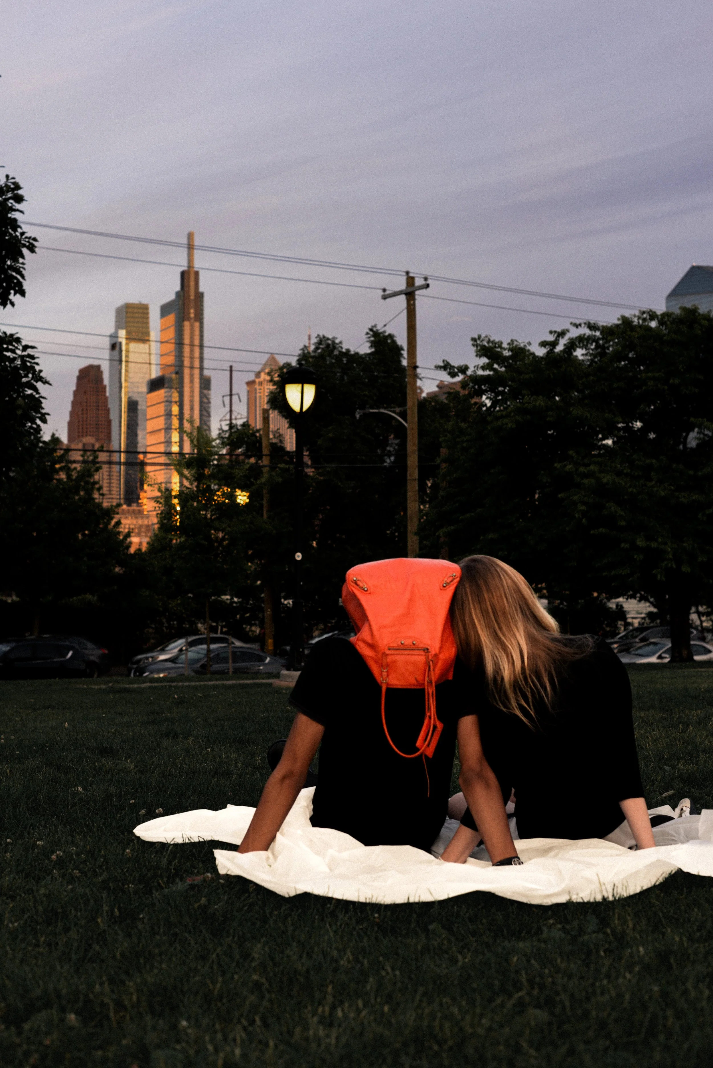 ; A couple sits on a picnic blanket in a city park with an orange Balenciaga bag used as a surreal romantic symbol.