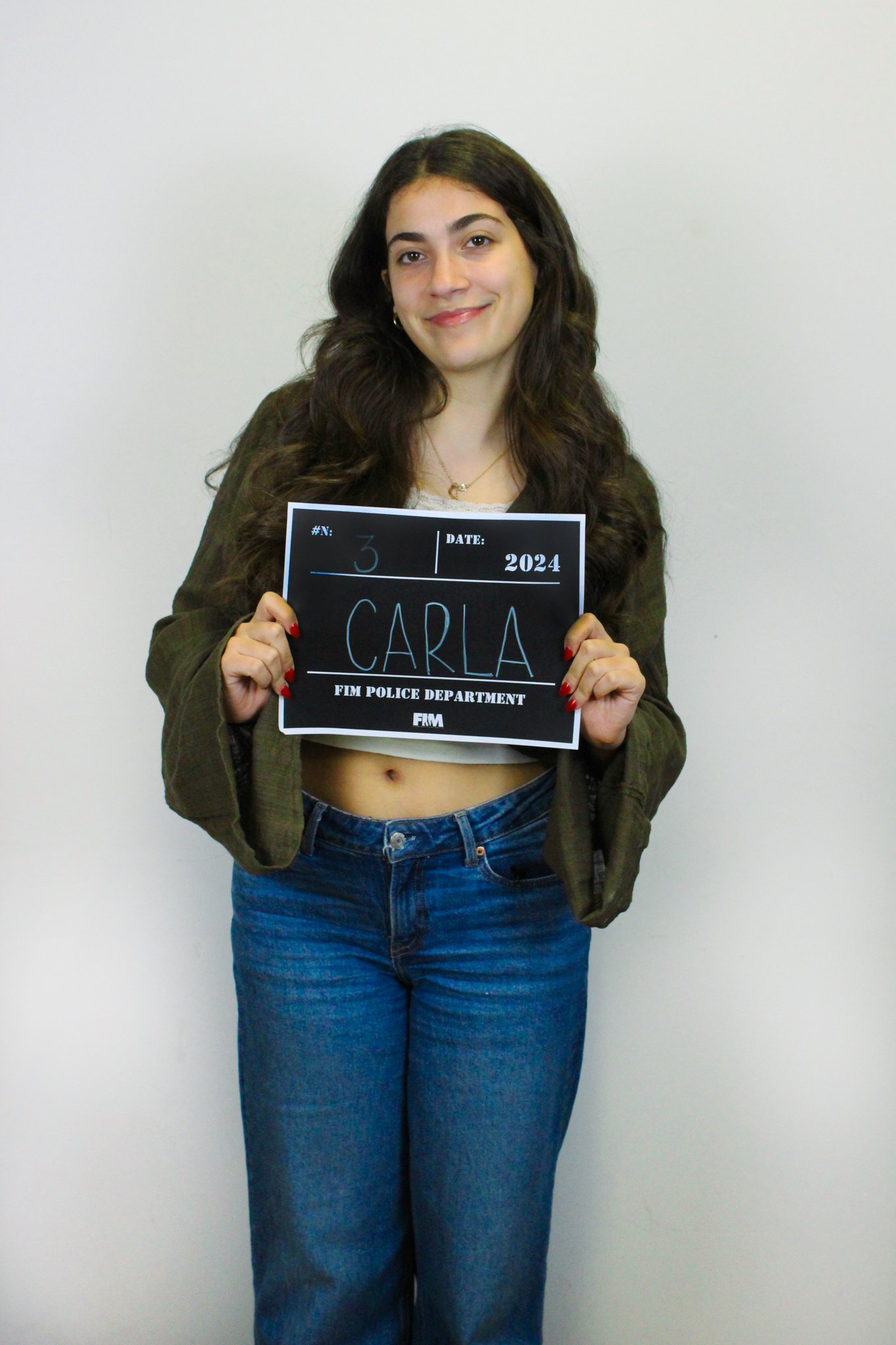 A woman with long dark hair, wearing a white crop top, jeans, and an olive green jacket, holds a sign with her name Carla and the date 2024, standing against a plain white wall.