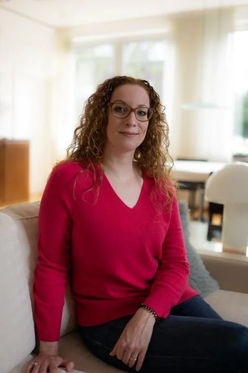 Frau mit Brille und lockigem Haar sitzt auf einem Sofa im Wohnzimmer.
