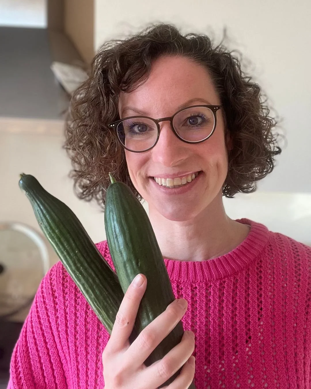 Eine lächelnde Frau mit Brille trägt einen pinkfarbenen Pullover und hält zwei Gurken in der Hand.