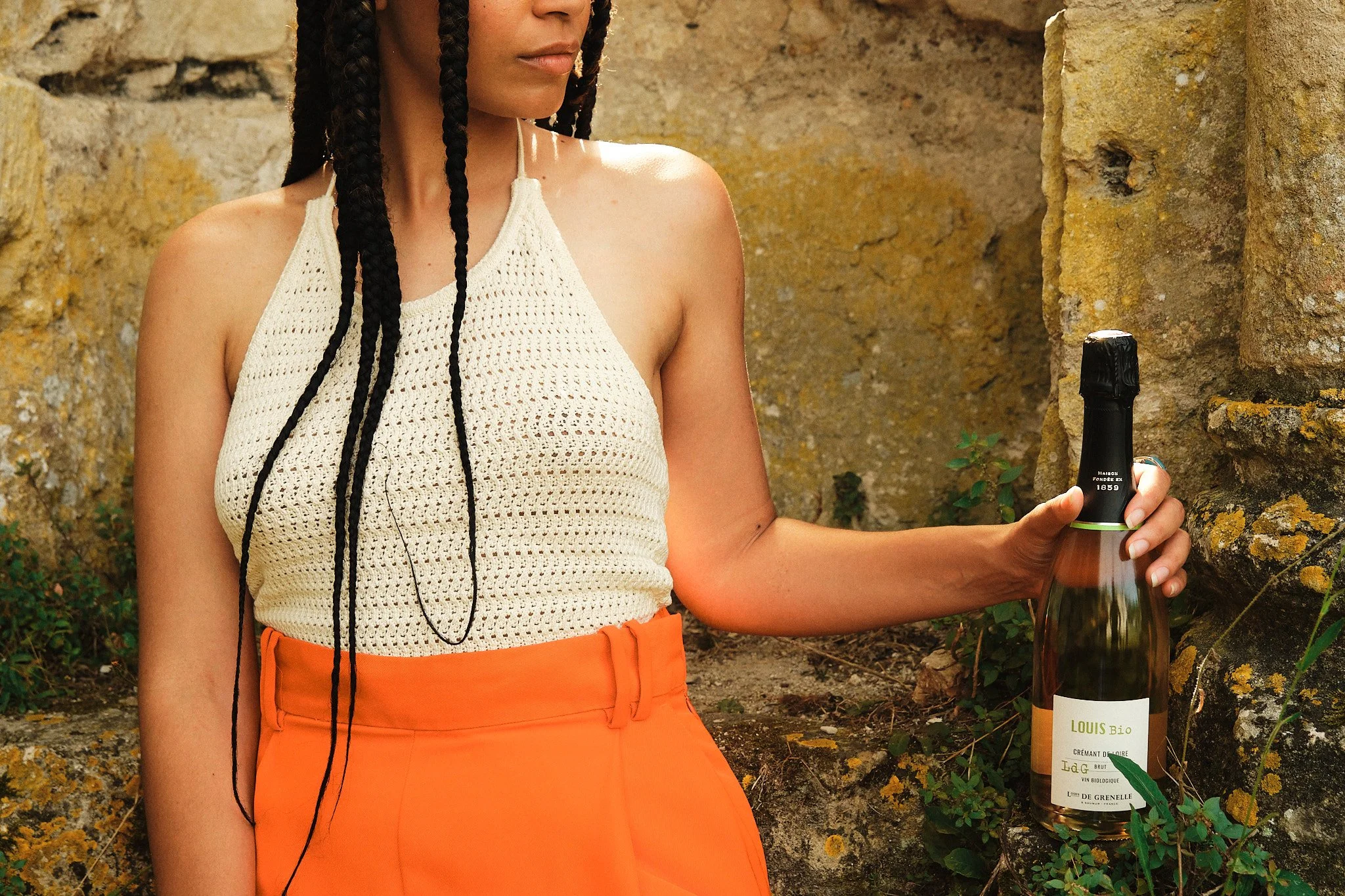 Une femme portant un pull blanc en crochet et une jupe orange tient une bouteille de champagne devant un mur en pierre.