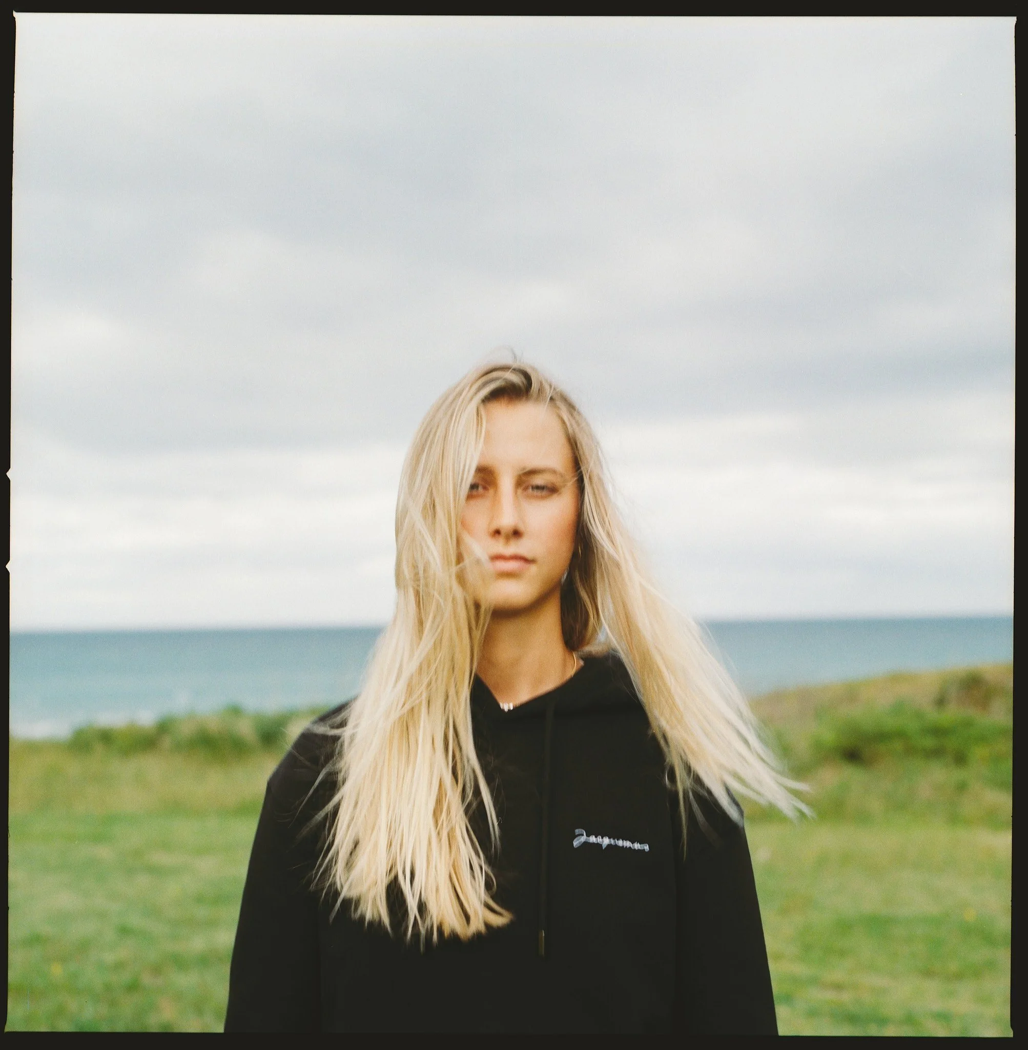 Jeune femme blonde portant un sweat à capuche noir, debout dans un paysage extérieur avec la mer et un ciel nuageux en arrière-plan.