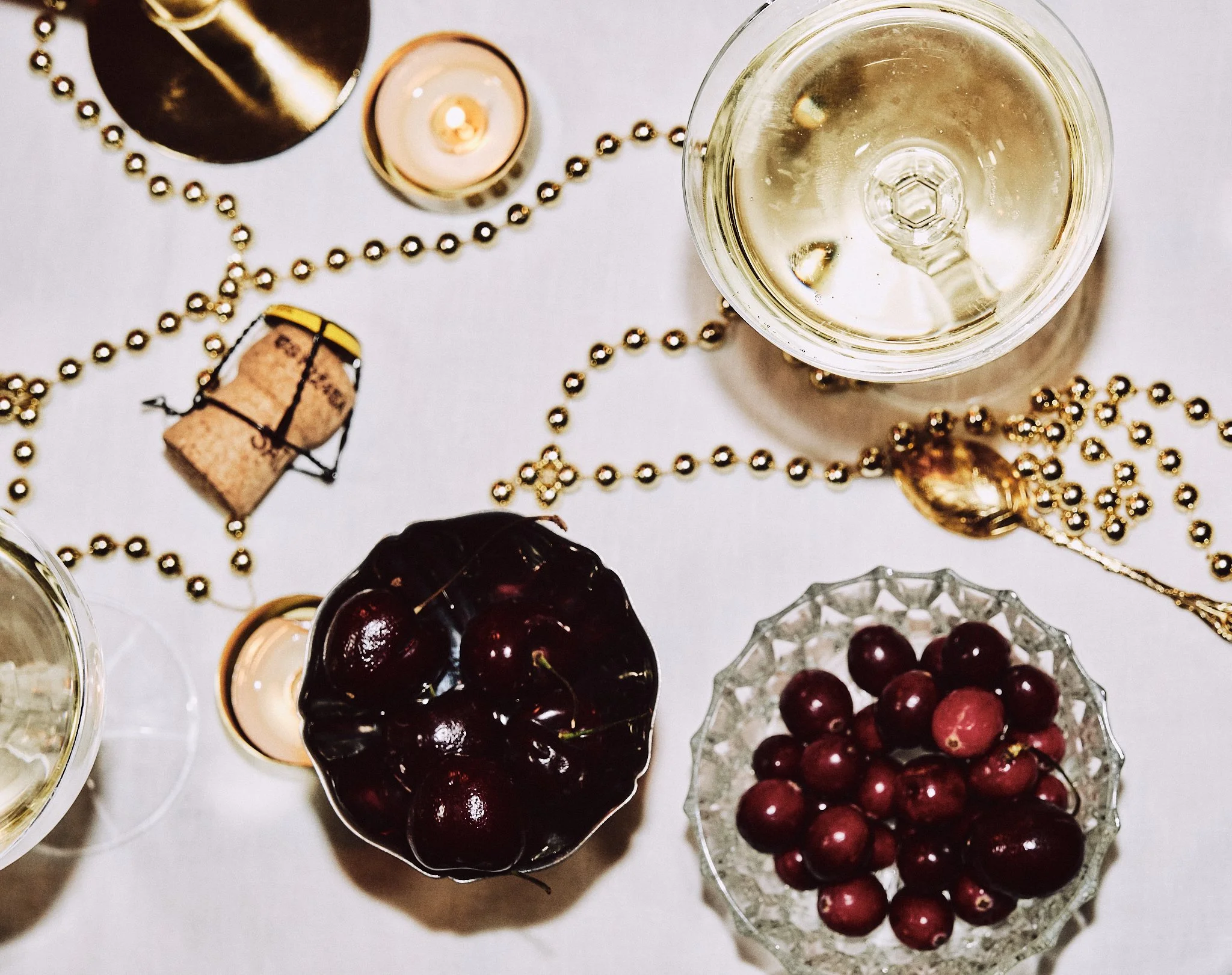 Verre de vin blanc, coupe de champagne, et assiettes de cerises rouges, avec des boules de perles dorées, bougie, et bouchon de champagne sur une table blanche.