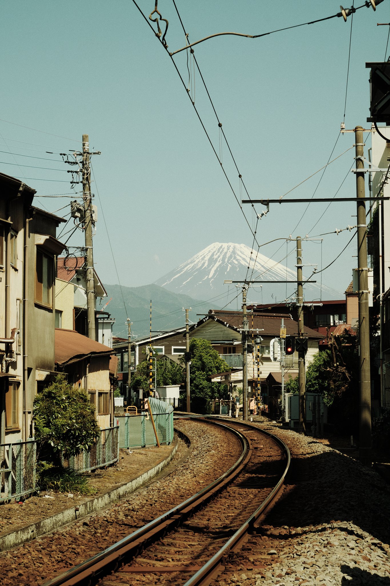 Une voie ferrée serpentante dans un quartier résidentiel avec la montagne du mont Fuji en arrière-plan, visible derrière les maisons et le réseau électrique.