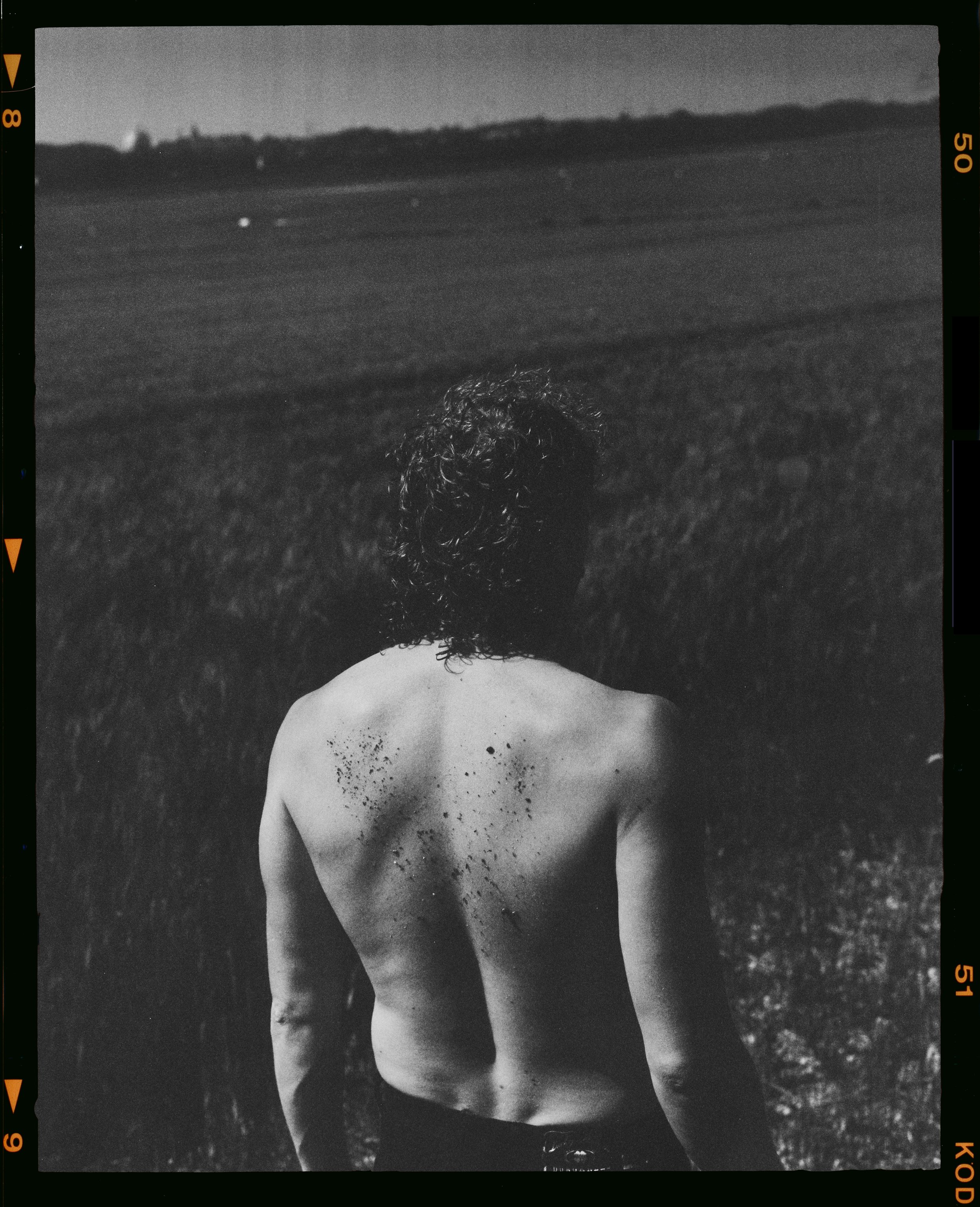 Une personne nue vue de dos, avec des cheveux bouclés, dans un champ en plein air, regardant l'horizon, en noir et blanc.