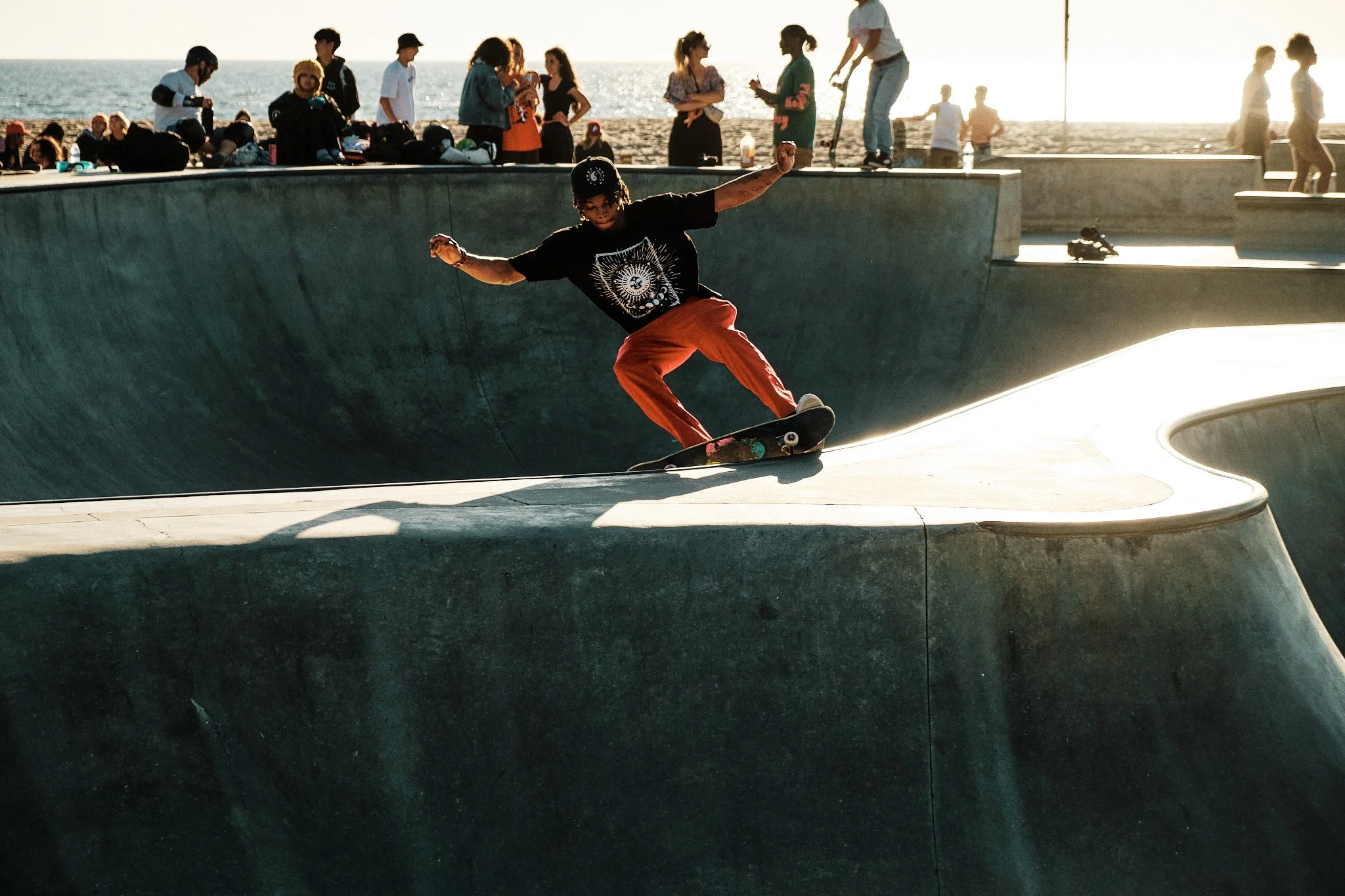 Jeune skateur faisant une figure dans un skatepark en bord de mer en fin d'après-midi. Beaucoup de spectateurs au fond, certains debout et d'autres assis, regardant les activités. Le soleil brille, créant des reflets sur la mer.