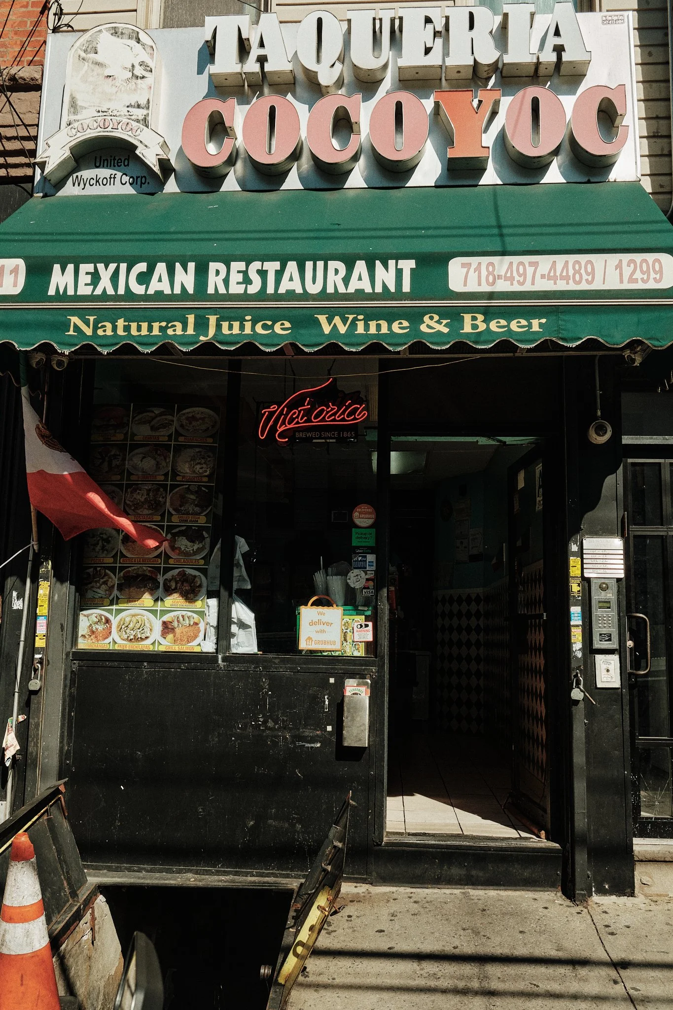 Façade d'un restaurant mexicain nommé Taqueria Cocoyoc, avec une enseigne verte, un panneau avec des photos de plats, une signalisation en néon rouge, et un bandeau mentionnant la livraison via Grubhub.