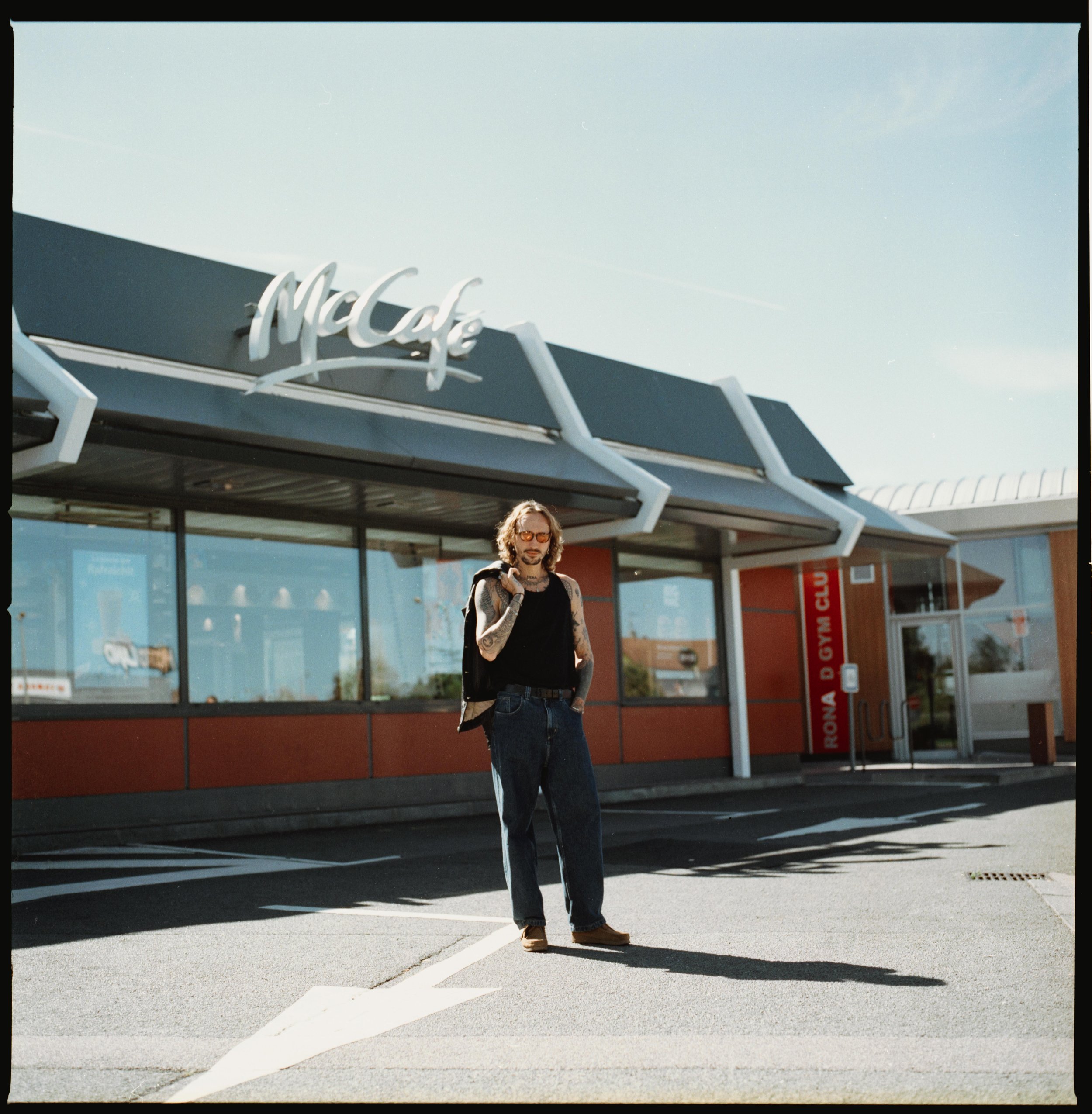 Un homme avec des tatouages, portant des lunettes de soleil, debout devant un restaurant McDonald's, sous un ciel ensoleillé.