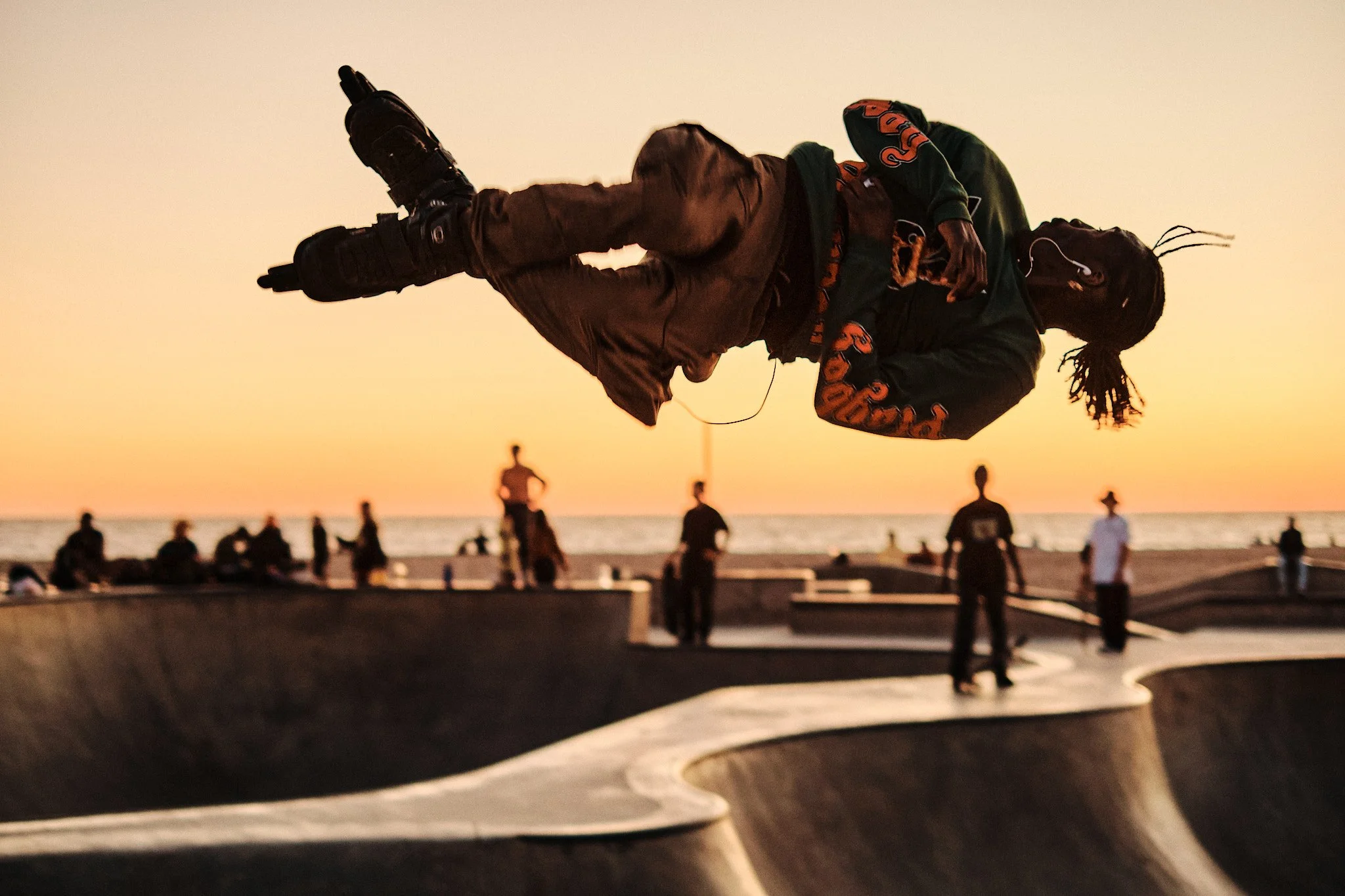 Un skateur effectuant un saut dans un skatepark au coucher du soleil, avec plusieurs personnes en arrière-plan et la mer en arrière-plan.