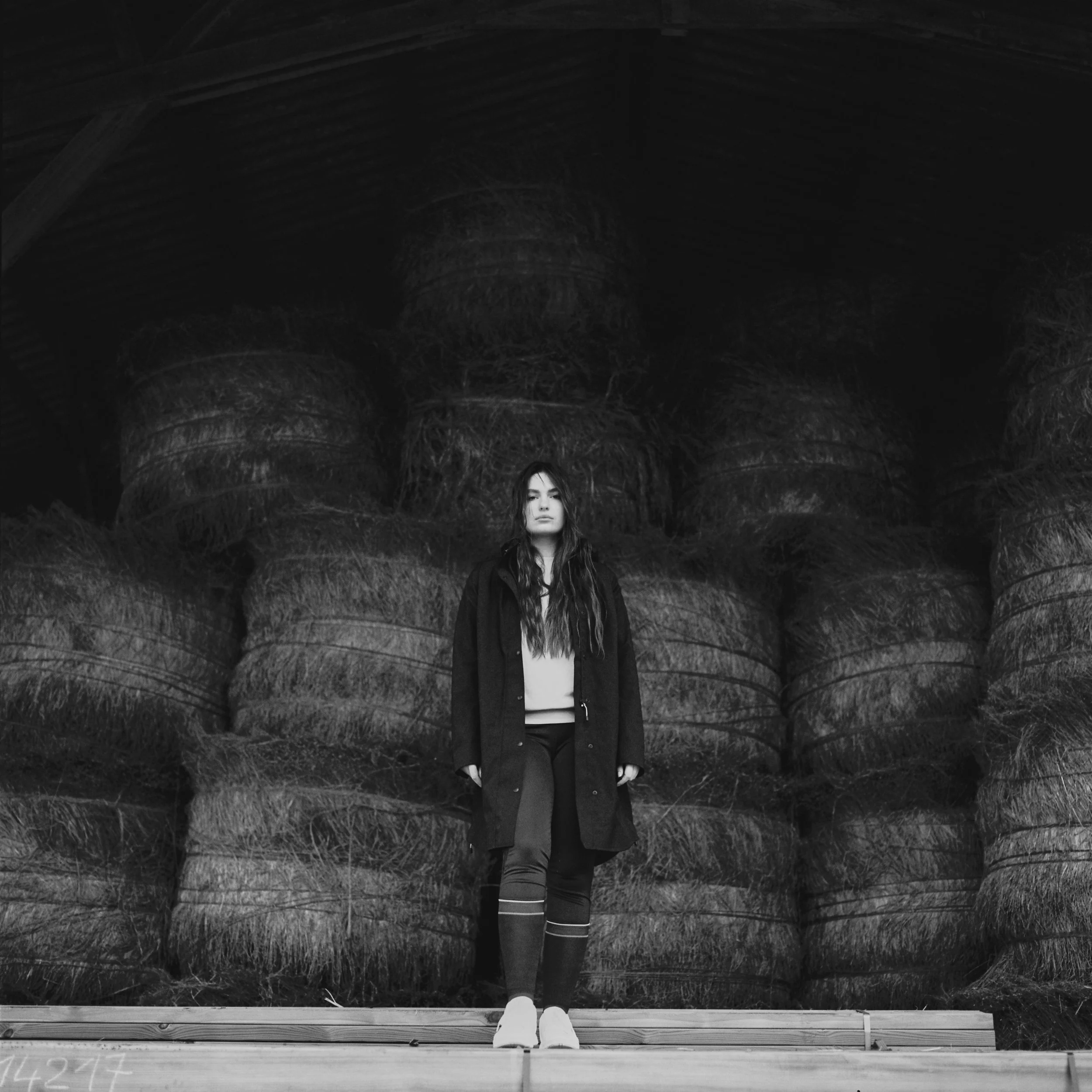 Une femme se tenant devant un grand tas de foin dans un bâtiment en bois, en noir et blanc.