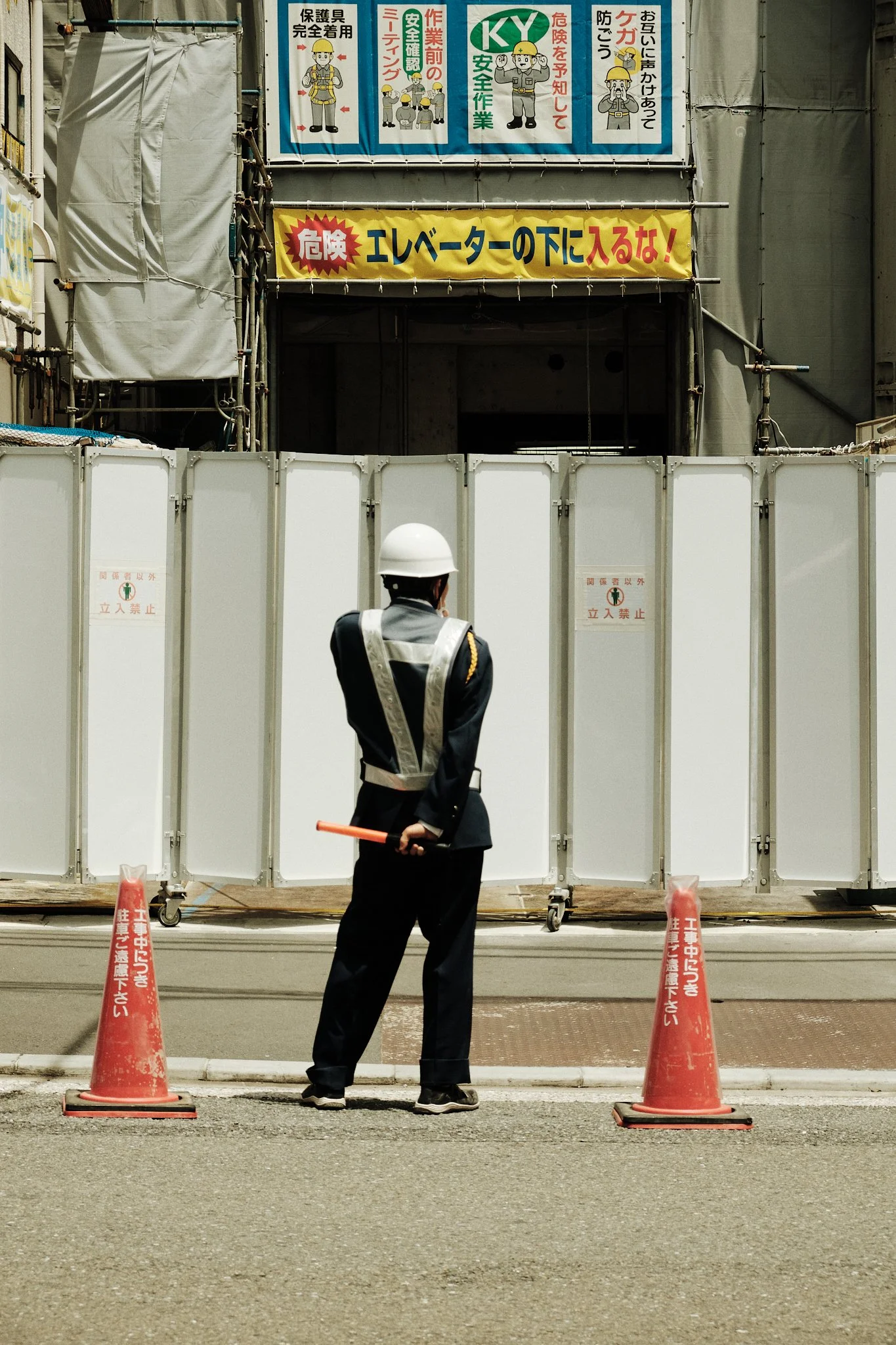 Un agent de sécurité en uniforme blanc et noir avec un casque, tenant une batte, se tient devant une barrière de sécurité blanche, dans une zone de construction au Japon. Il y a deux cônes orange avec des inscriptions en japonais sur le trottoir.