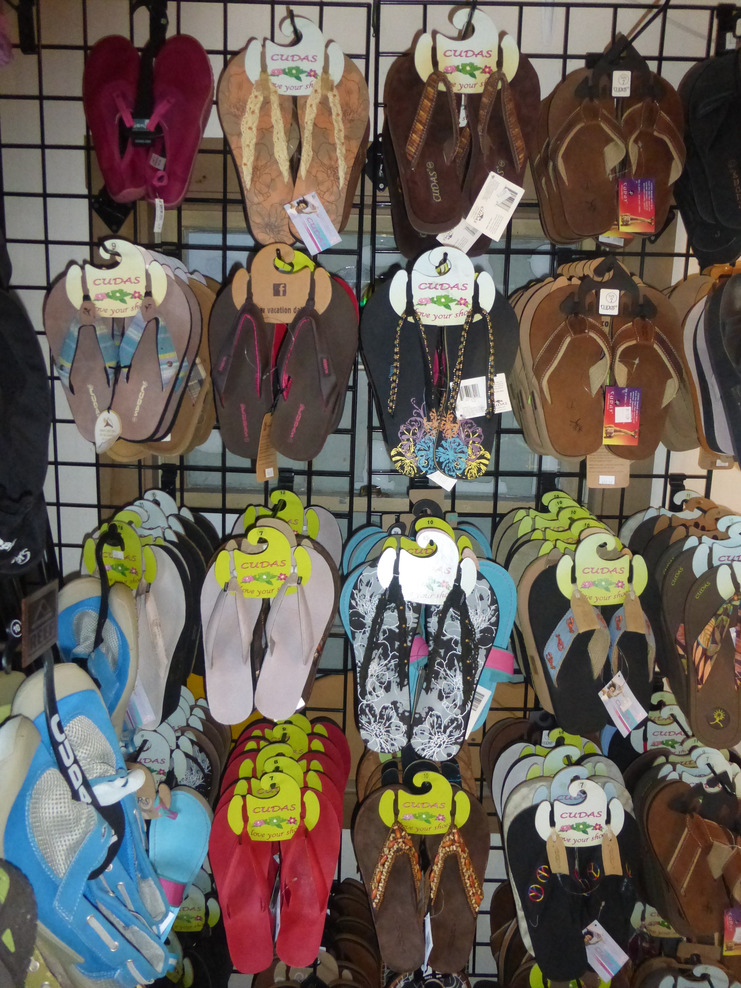 Display of various flip-flops from the brand Cudas hanging on a grid wall. Multiple colors and styles are visible, including plain and patterned designs.
