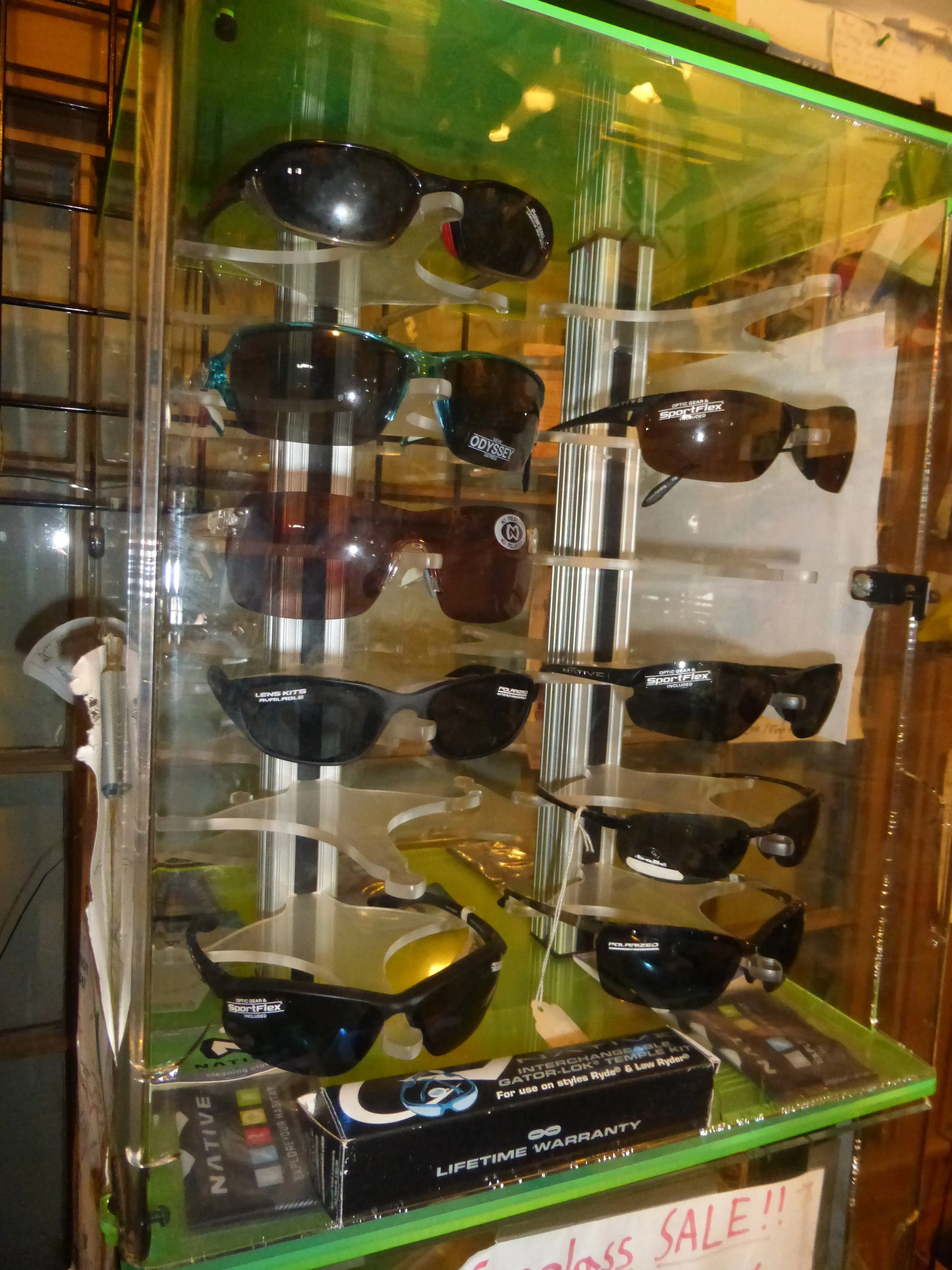 Display case with various sunglasses, including brands like Spot Flex and Odyssey, on a green shelf with a lifetime warranty promotion.