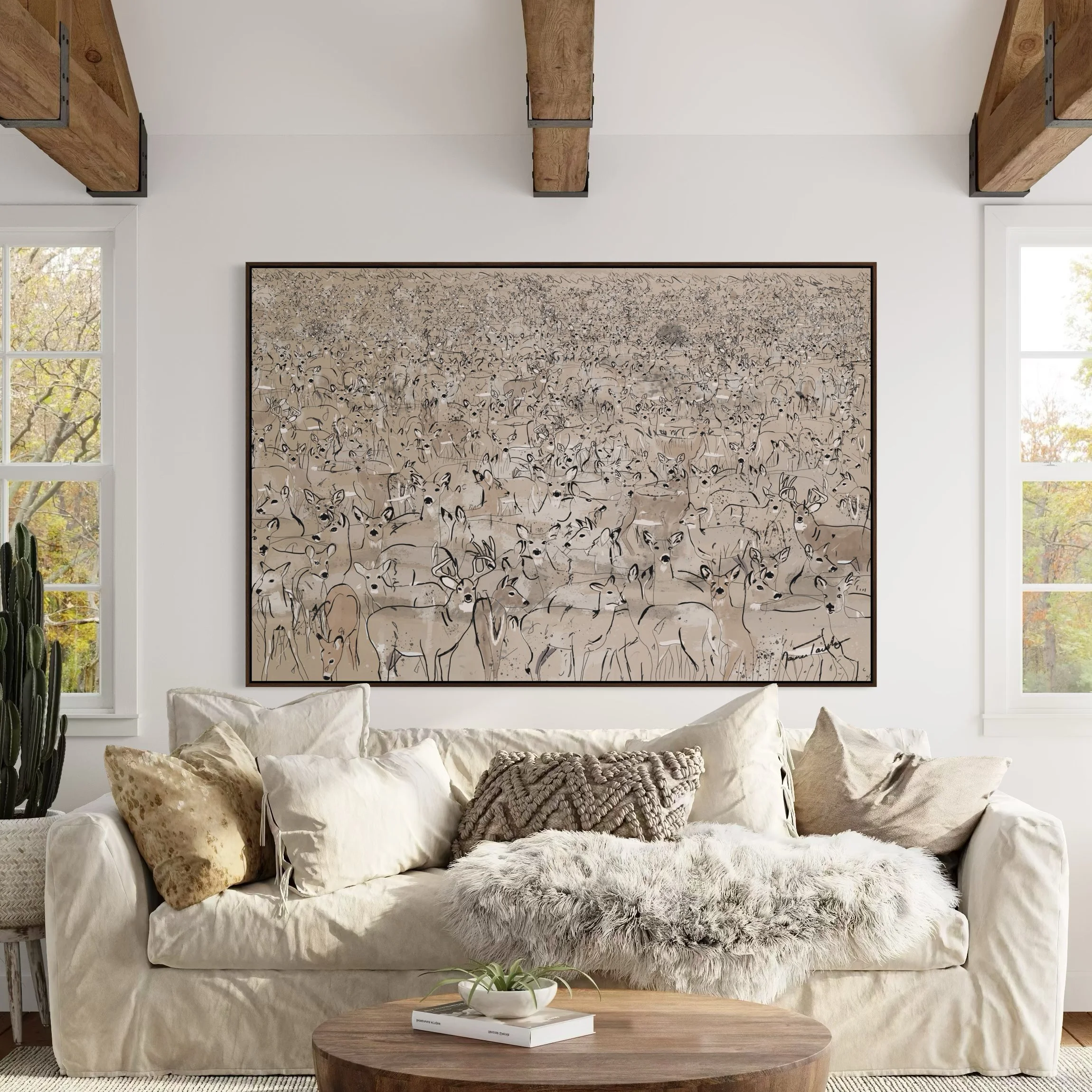 Living room with white sofa, patterned pillows, glass coffee table with plant, large framed artwork of a herd of deer, white walls, windows with trees outside, exposed wooden beams.